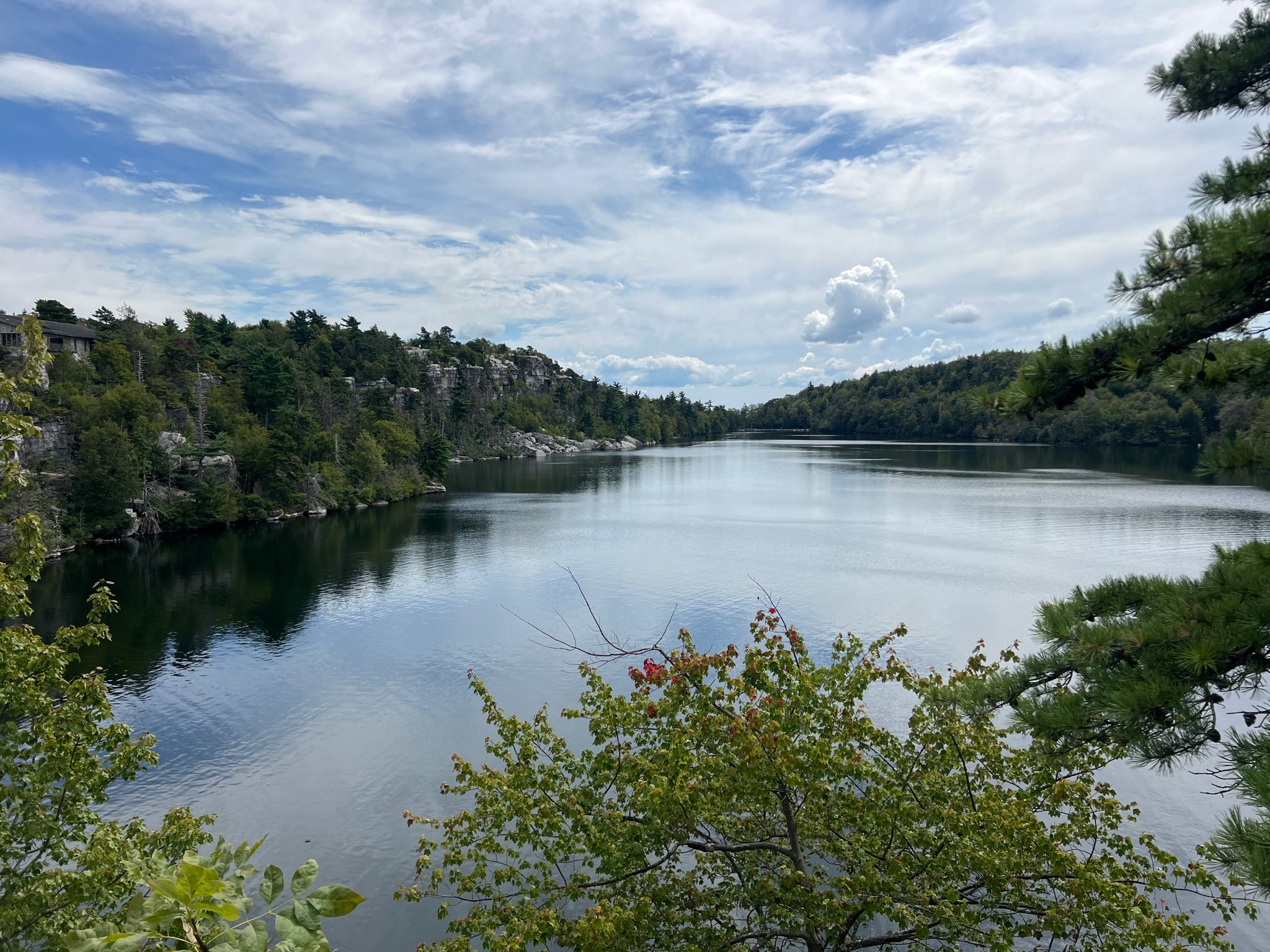 Calm lake reflecting a partly cloudy sky, surrounded by green forested hills. - Lake Minnewaska Carriage Road Loop