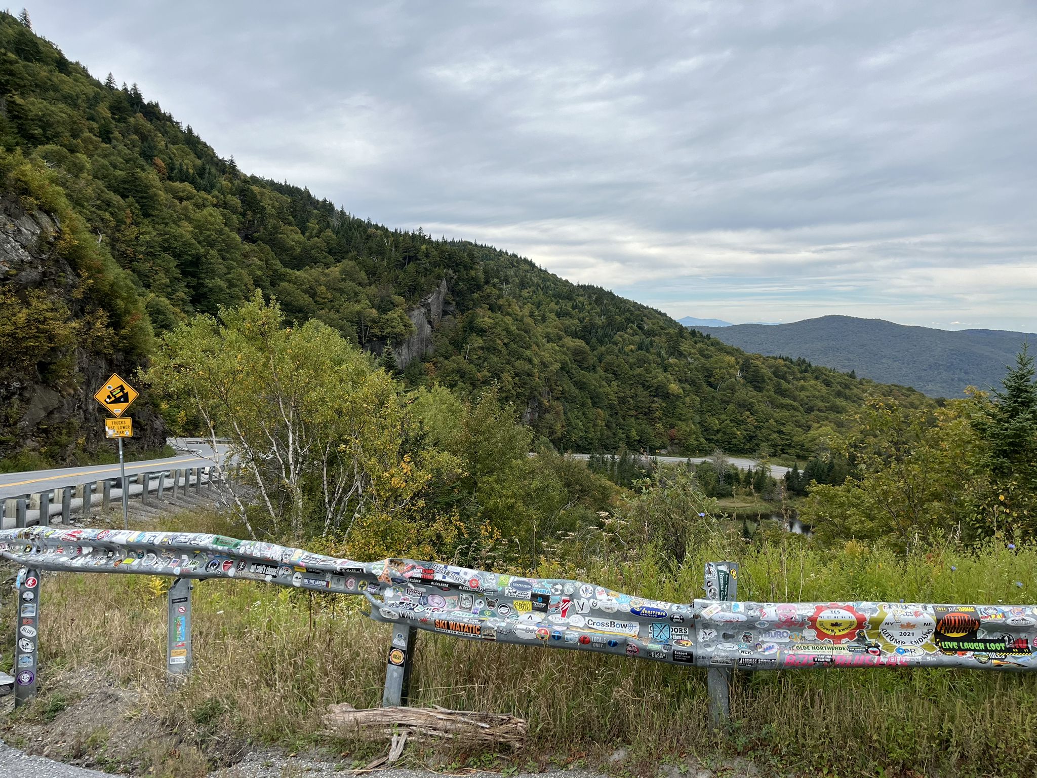 Sticker-covered guardrail beside a winding mountain road on an overcast day. - Middlebury College Quad – Middlebury Falls loop from Middlebury