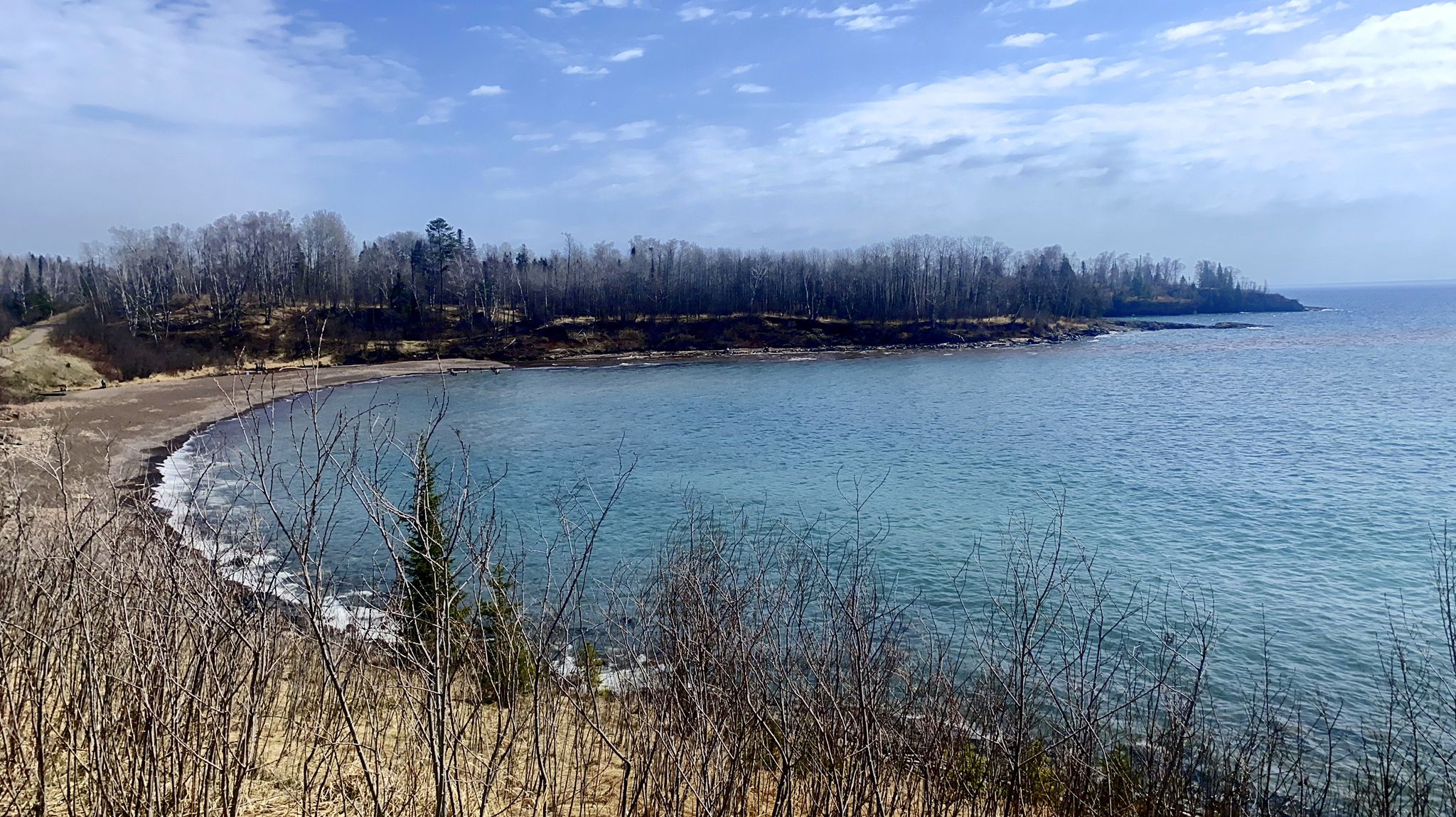 A calm blue bay with a rocky beach and bare trees under a bright sky. - Gitchi-Gami State Trail