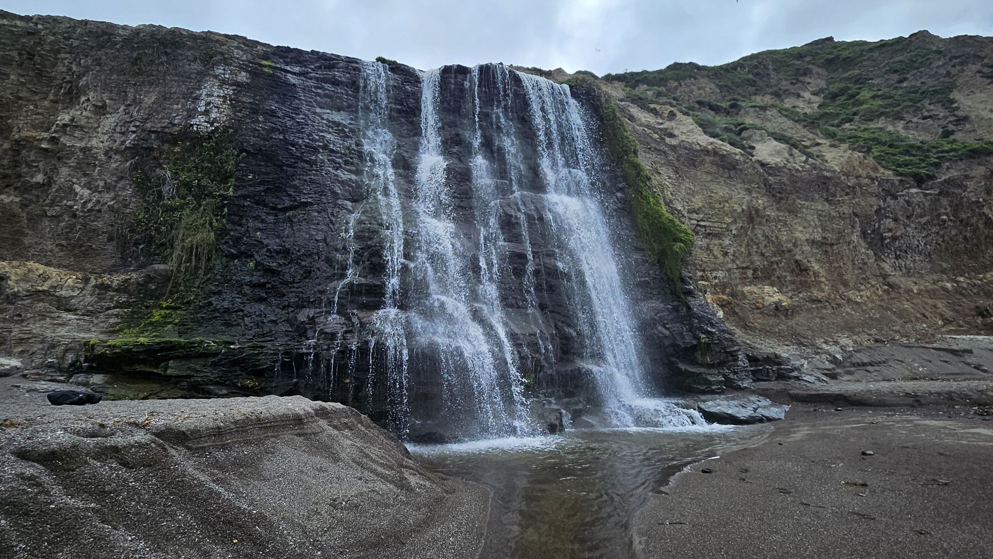Large waterfall cascading down a dark, mossy cliff onto a sandy beach. - Stewart Trail Long Loop