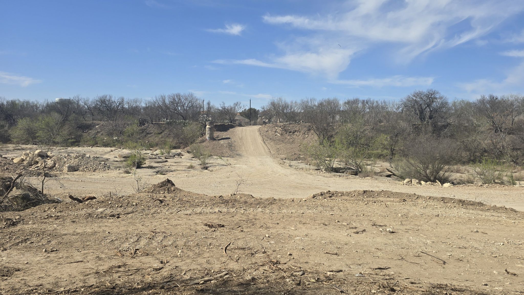 A wide dirt road leads uphill through dry, sparse trees under a blue sky. - Leon Creek Greenway Trail