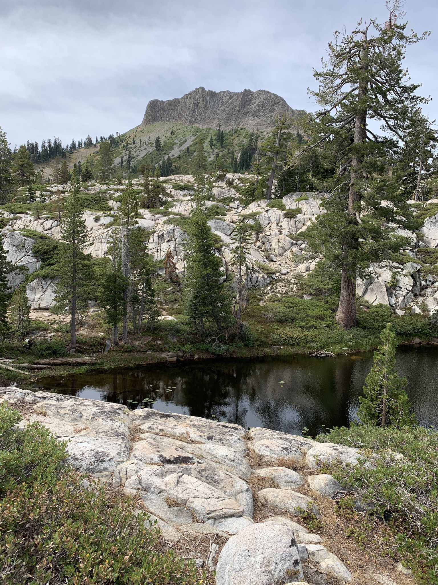 A dark lake reflecting a rugged mountain peak and rocky, forested landscape. - Palisades Creek Trail
