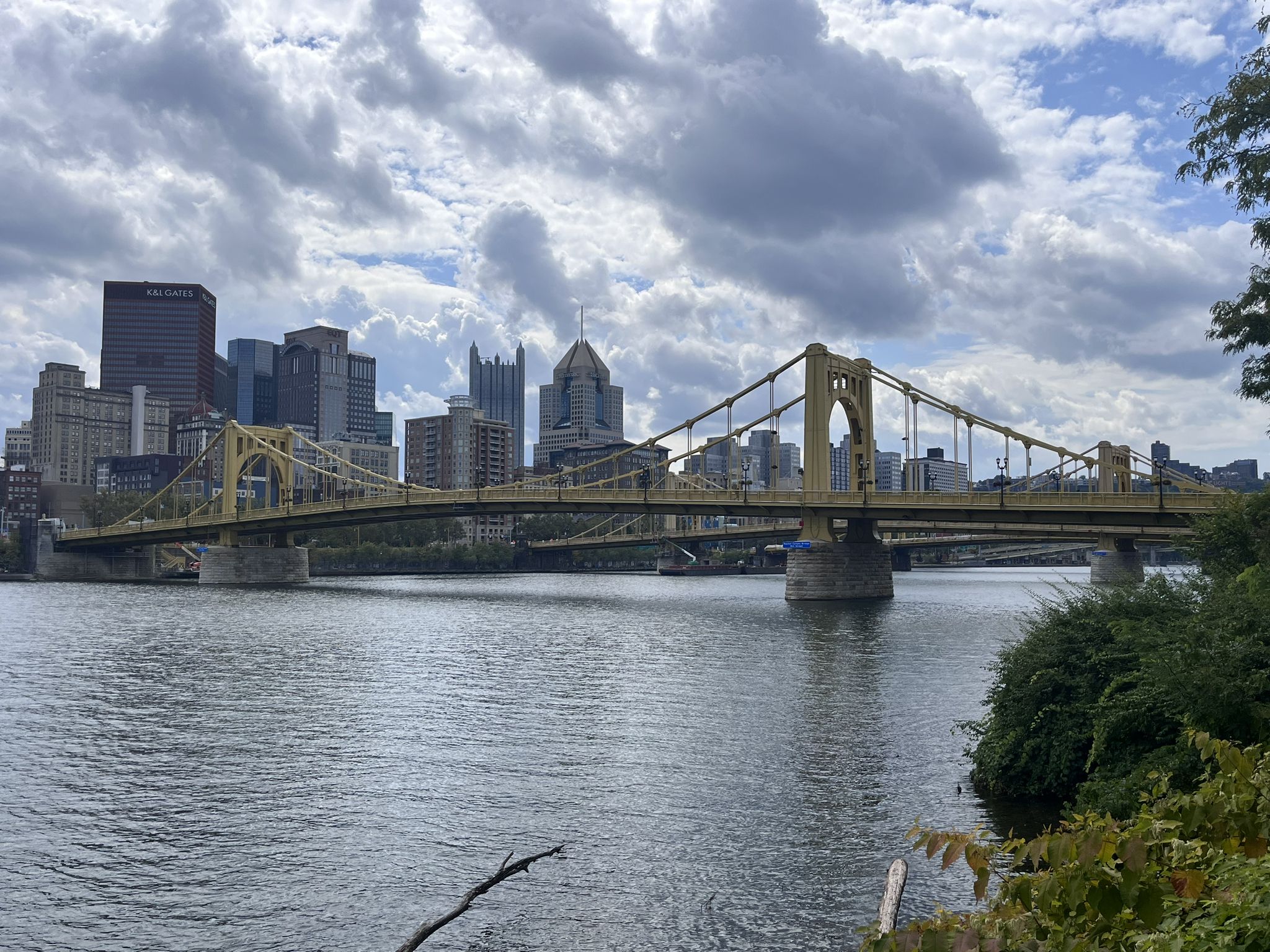 Yellow suspension bridge over a river with a city skyline under cloudy skies. - North Shore Trail