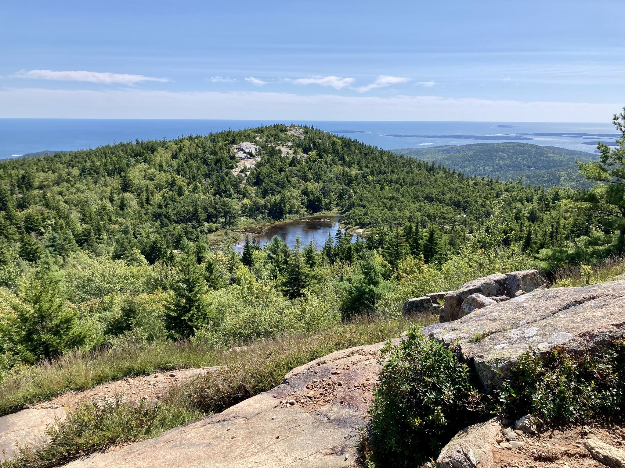 Mountain vista with a small lake, green forest, and distant ocean under blue sky. - Cadillac South Ridge Trail