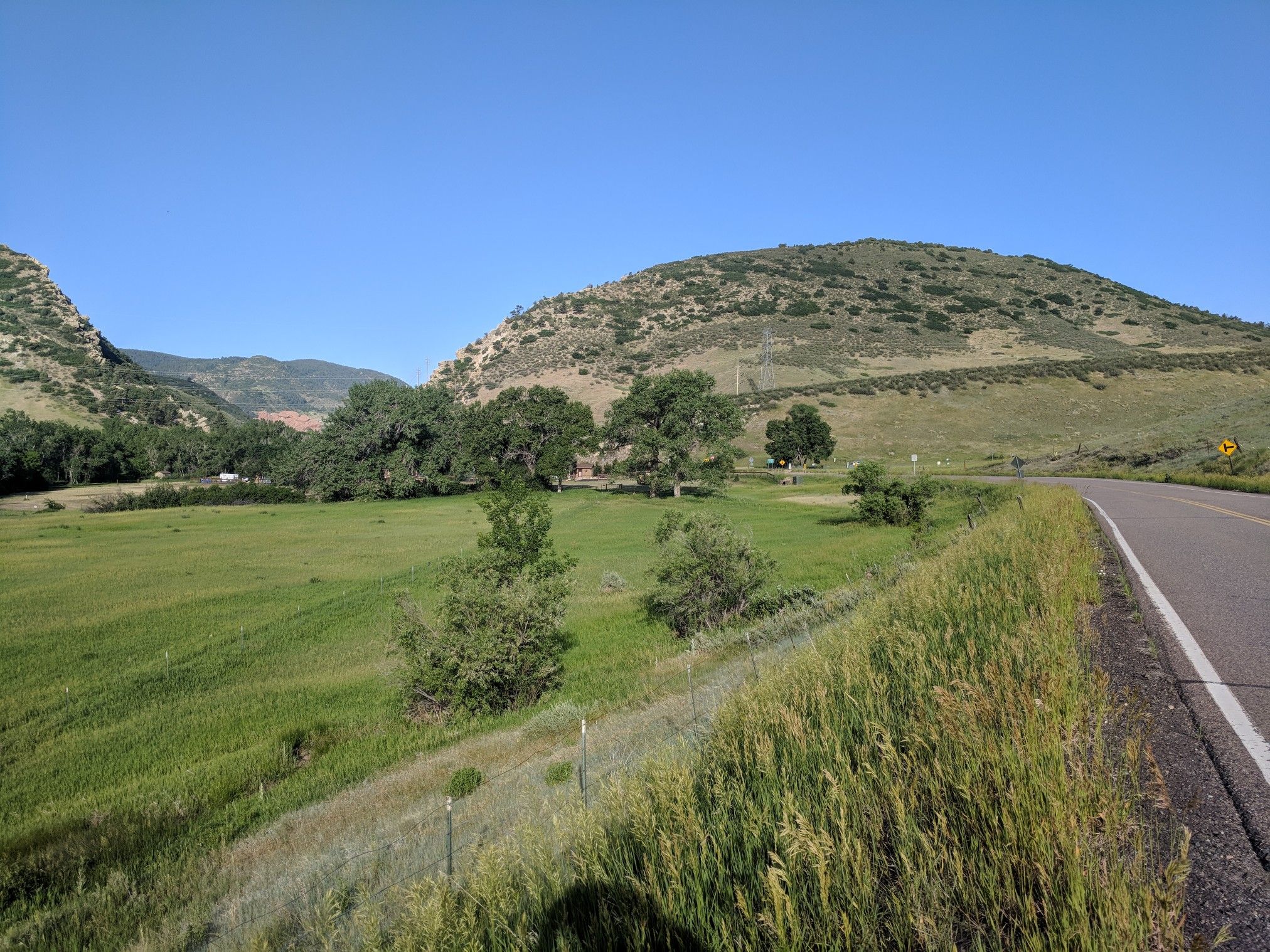 Paved road alongside a vibrant green field and rolling hills under a clear blue sky. - Pleasant Park Grange Rest Stop loop from Chatfield State Park