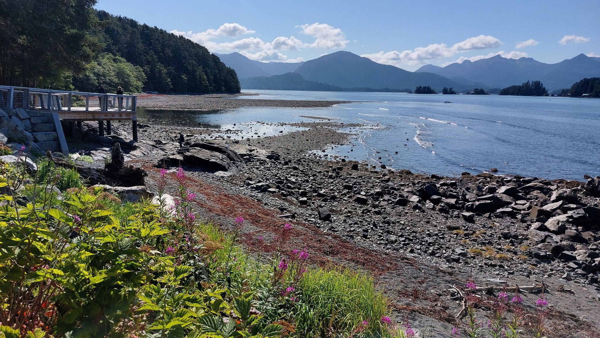 Rocky shoreline with calm water, distant mountains, and a wooden platform under sunny skies. - Sea Walk and Totem Trail Loop