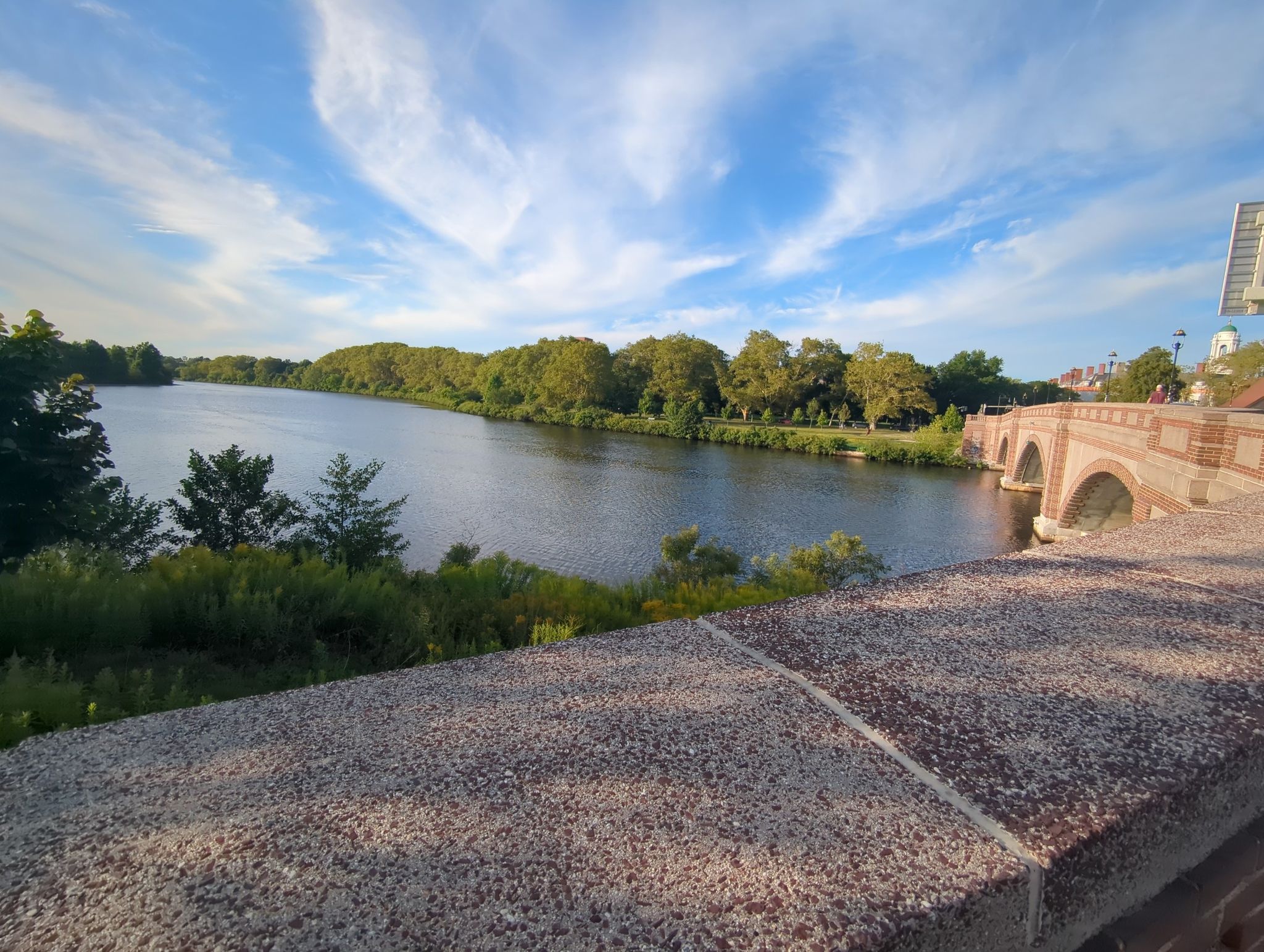 Wide river with a stone bridge and trees under a bright blue sky. - Charles River Greenway Loop