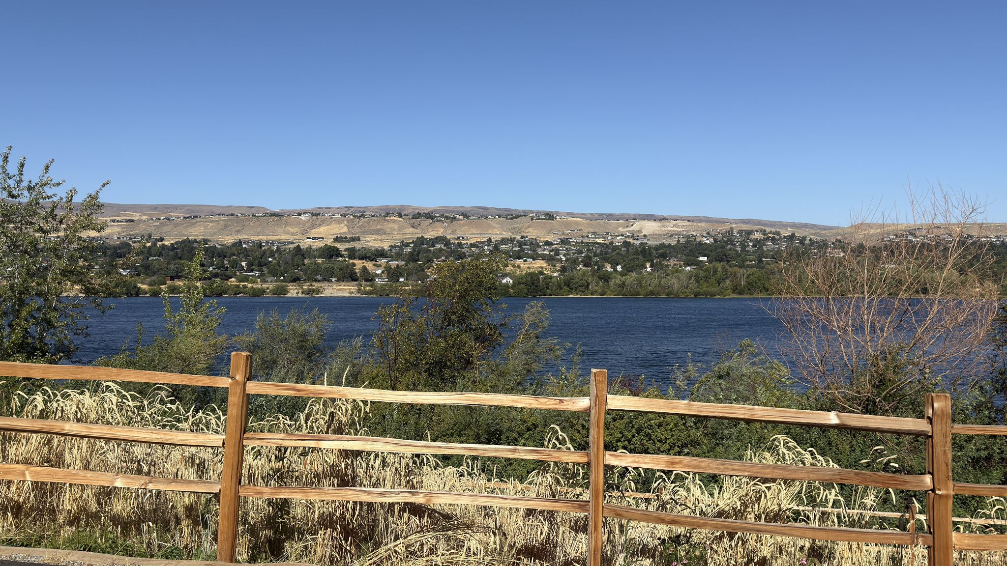 Wooden fence overlooking a wide lake and distant town, clear blue sky. - Walla Walla Point Park loop from Wenatchee