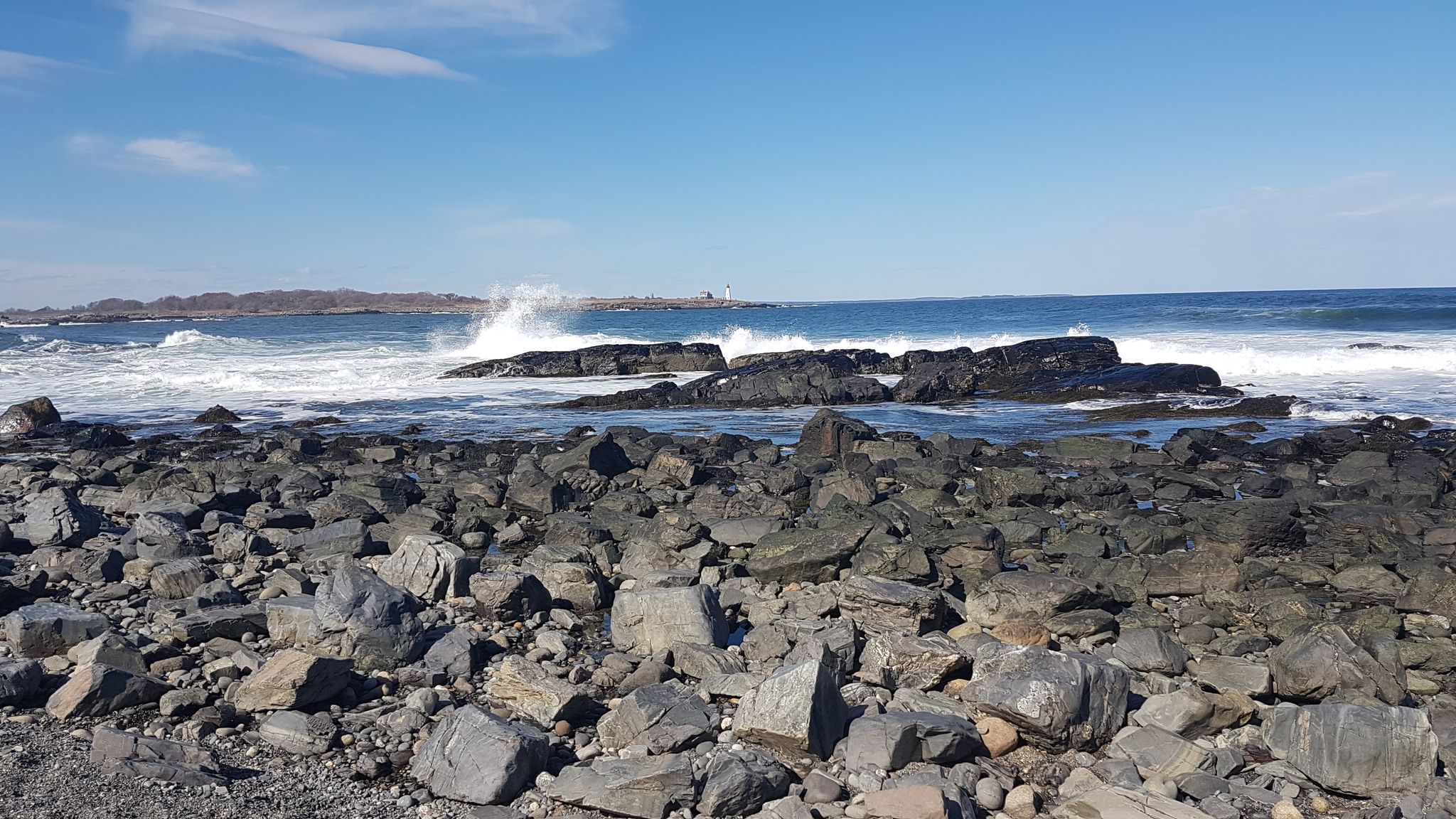 Large waves crash against dark rocks on a rugged, rocky beach under a blue sky. - East Point Audubon Sanctuary Trail
