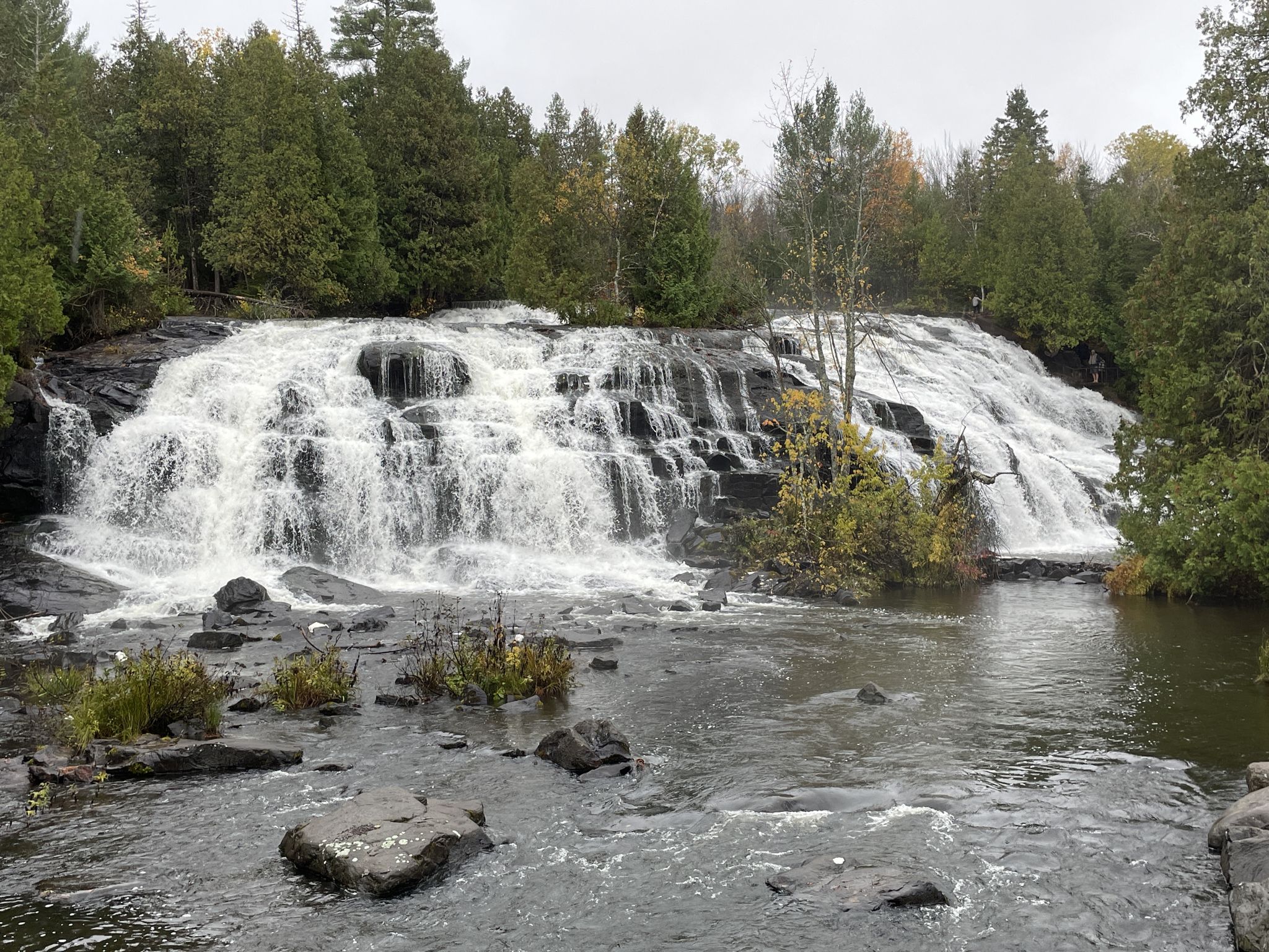Multi-tiered waterfall flows into a rocky river, surrounded by green and autumn trees. - Bond Falls Loop