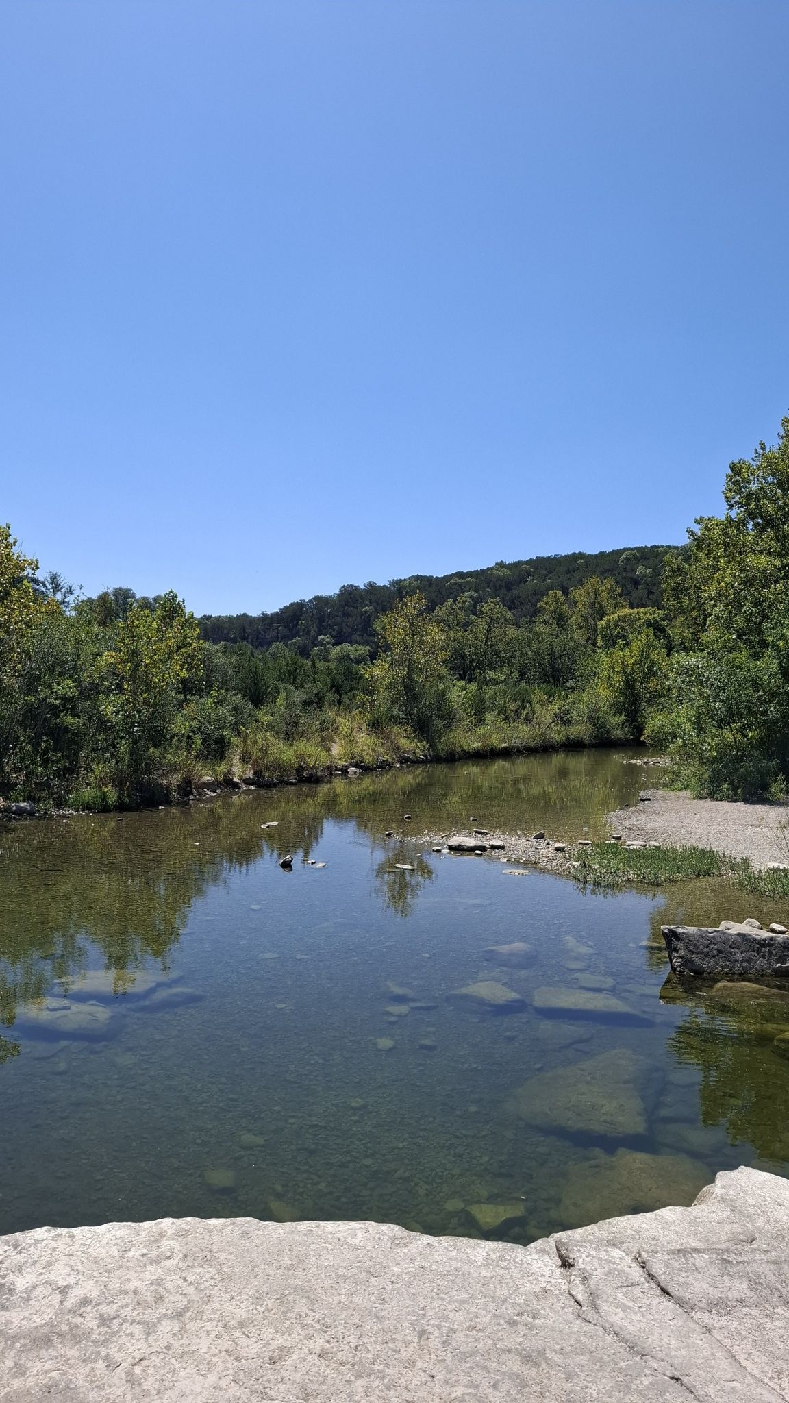 Calm river reflecting green trees and hills under a clear blue sky. - Barton Creek Greenbelt Trail