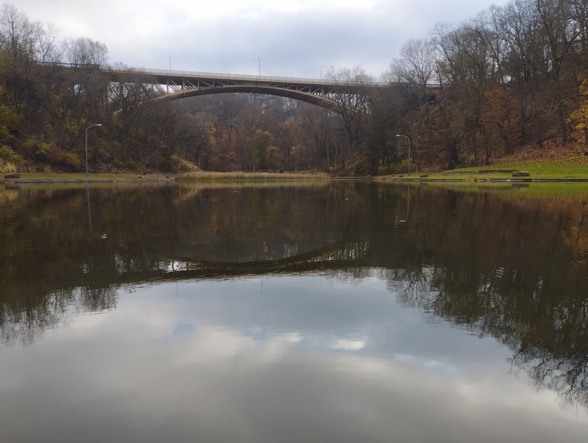 Calm pond reflecting a large arch bridge and cloudy sky, surrounded by trees. - Schenley Park loop via Hollow Run Trail
