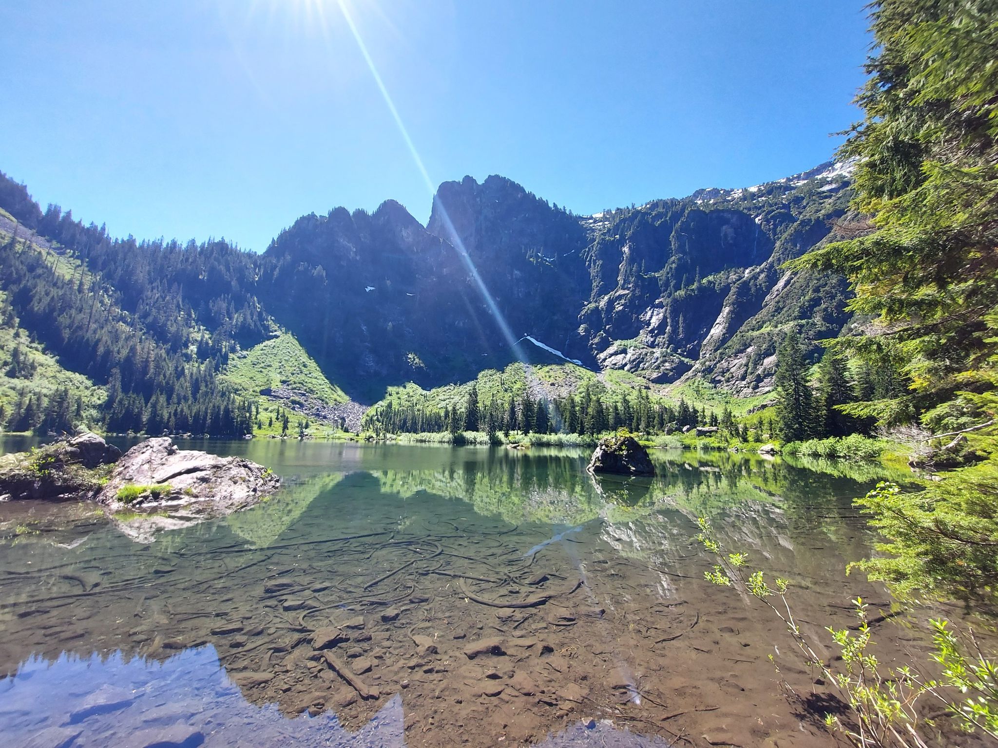 Clear mountain lake reflecting rugged peaks and green forest on a sunny day. - Heather Lake Trail