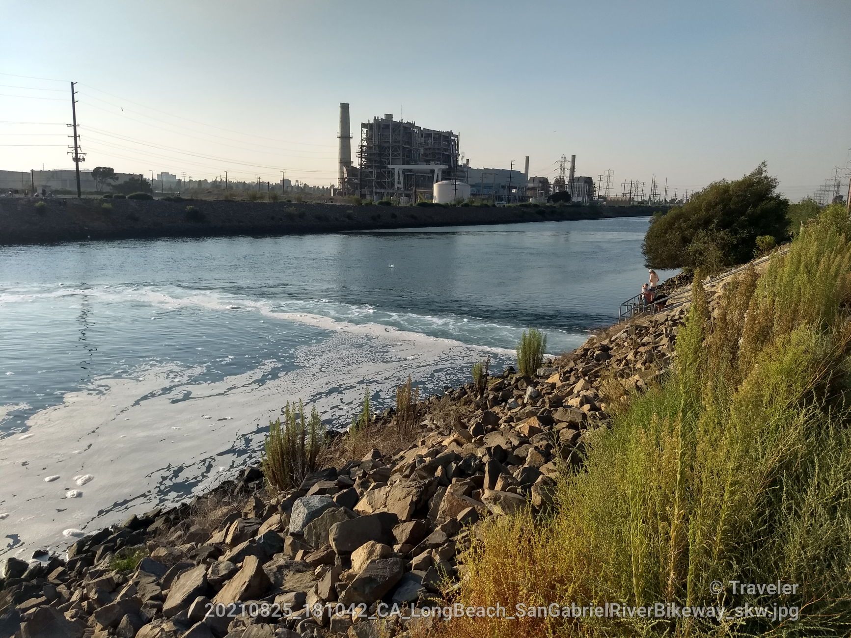 River with foamy water, rocky bank, and distant industrial buildings under clear sky. - San Gabriel River Trail – San Gabriel River Trail loop from Seal Beach National Wildlife Refuge