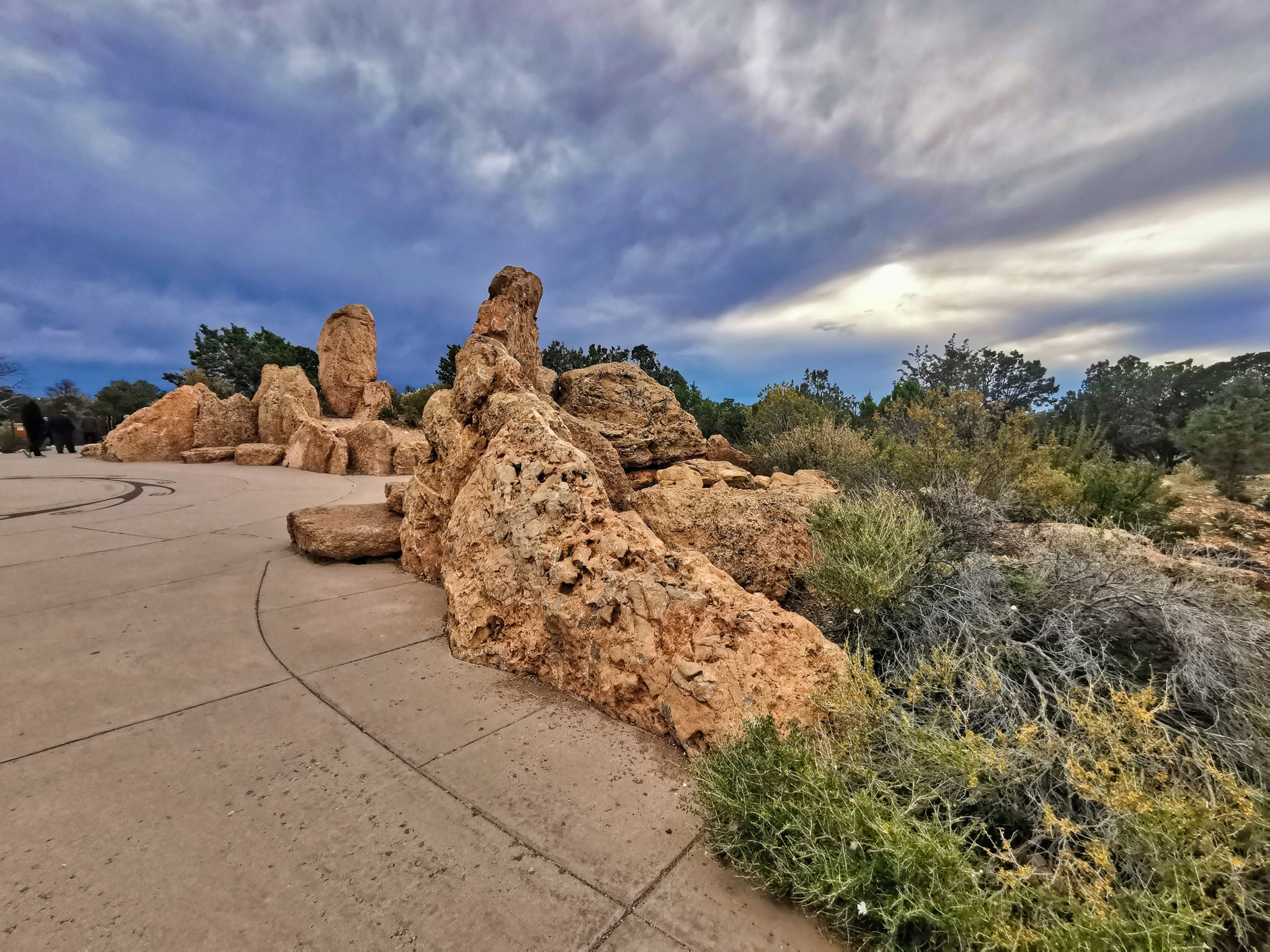 Large rock formations and green bushes next to a paved path under cloudy sky. - Rim Trail: Mather Point to Yavapai Point