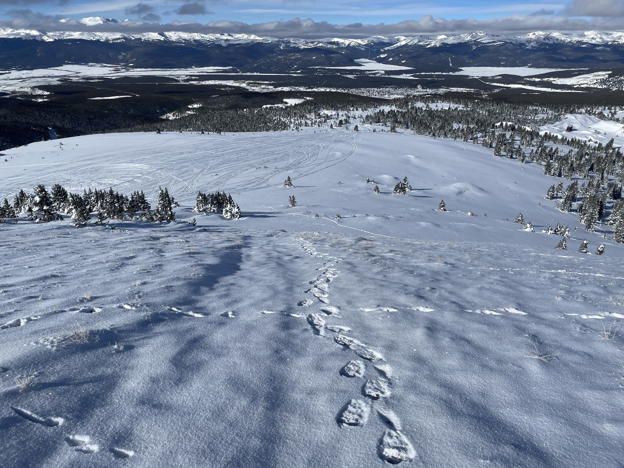 Footprints lead across a sunny, snow-covered hill with distant mountains and trees. - Ball Mountain loop
