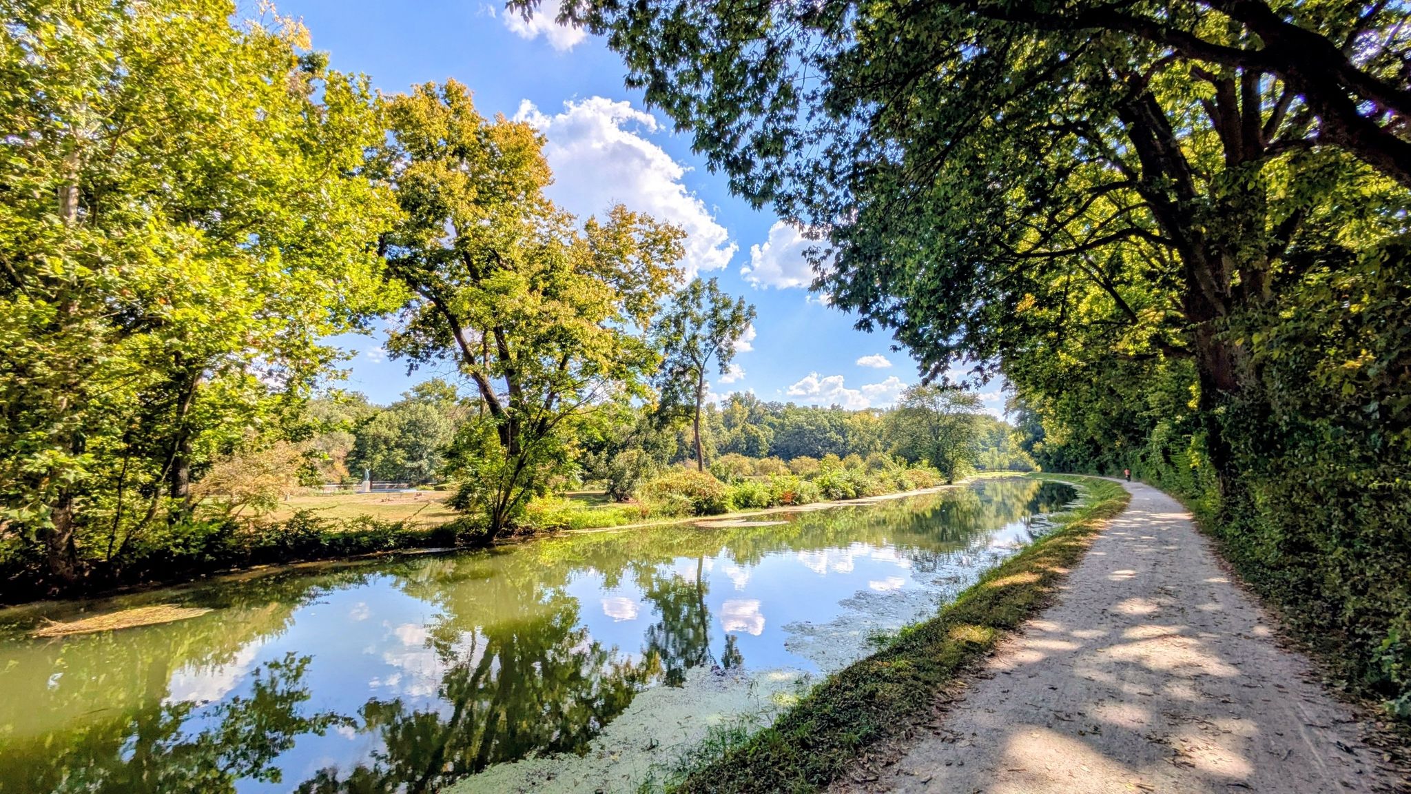 Sunny canal with calm water reflecting trees and sky, alongside a dirt path. - Butler University – Butler University loop from Butler University