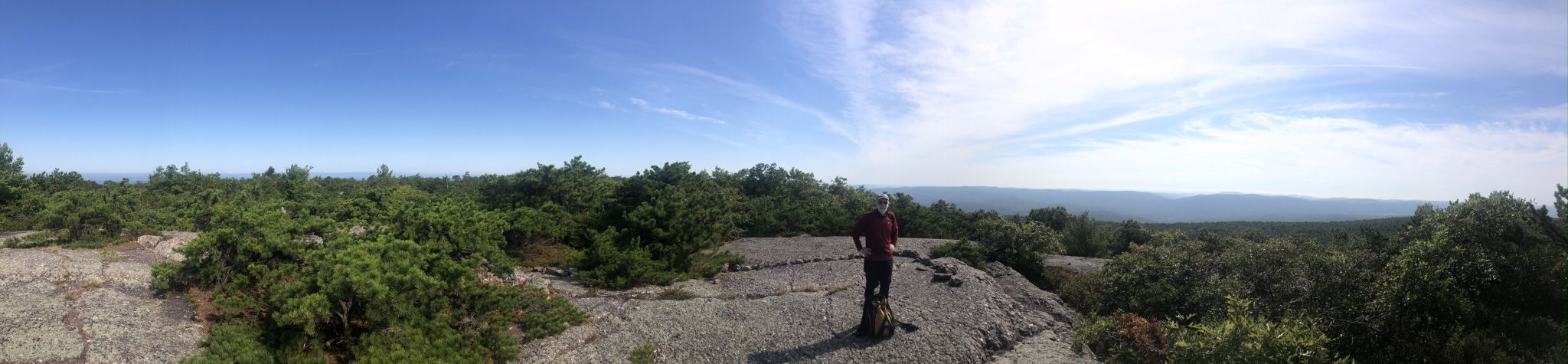 A person stands on a rocky overlook, enjoying a vast sunny forested panorama. - Schunnemunk Mountain via Jessup Trail