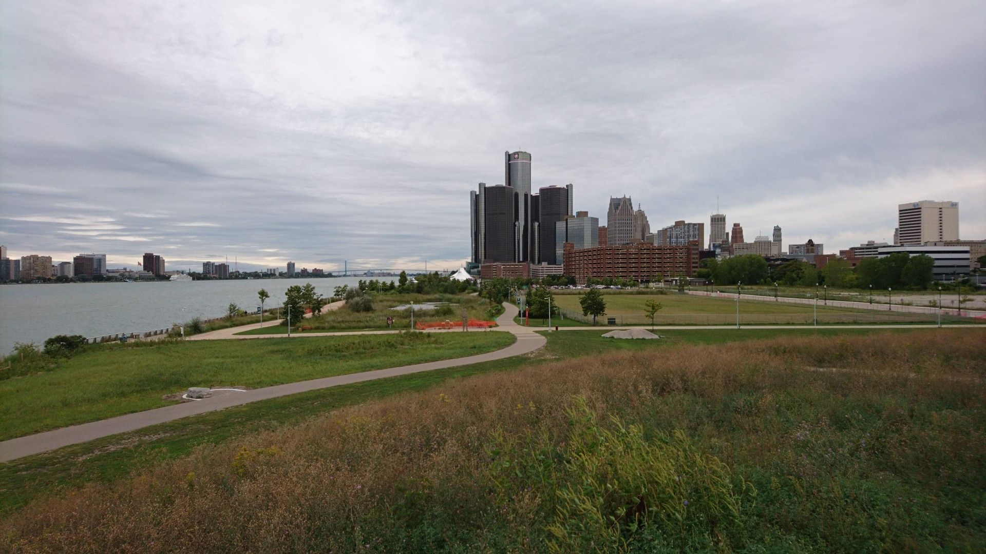 City skyline across a wide river with green parkland under cloudy sky. - Detroit Riverwalk Viewpoint – William G. Milliken State Park loop from Detroit
