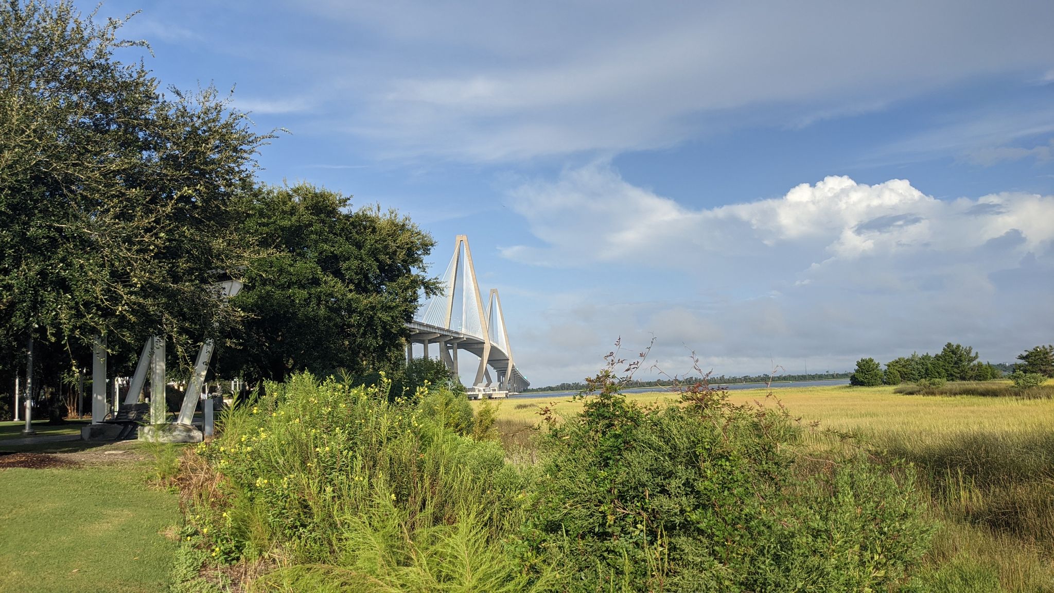 Lush green foliage framing a large white bridge over water under a cloudy sky. - Patriots Point Loop