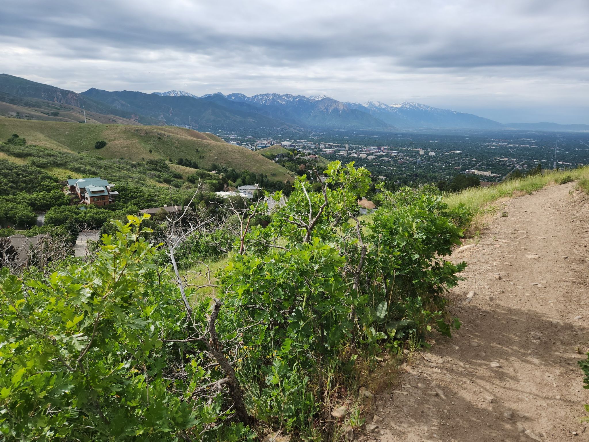 Dirt path, green bushes, distant city, and snowy mountains under cloudy sky. - Great view of Salt Lake City – University of Utah Campus loop from Fort Douglas