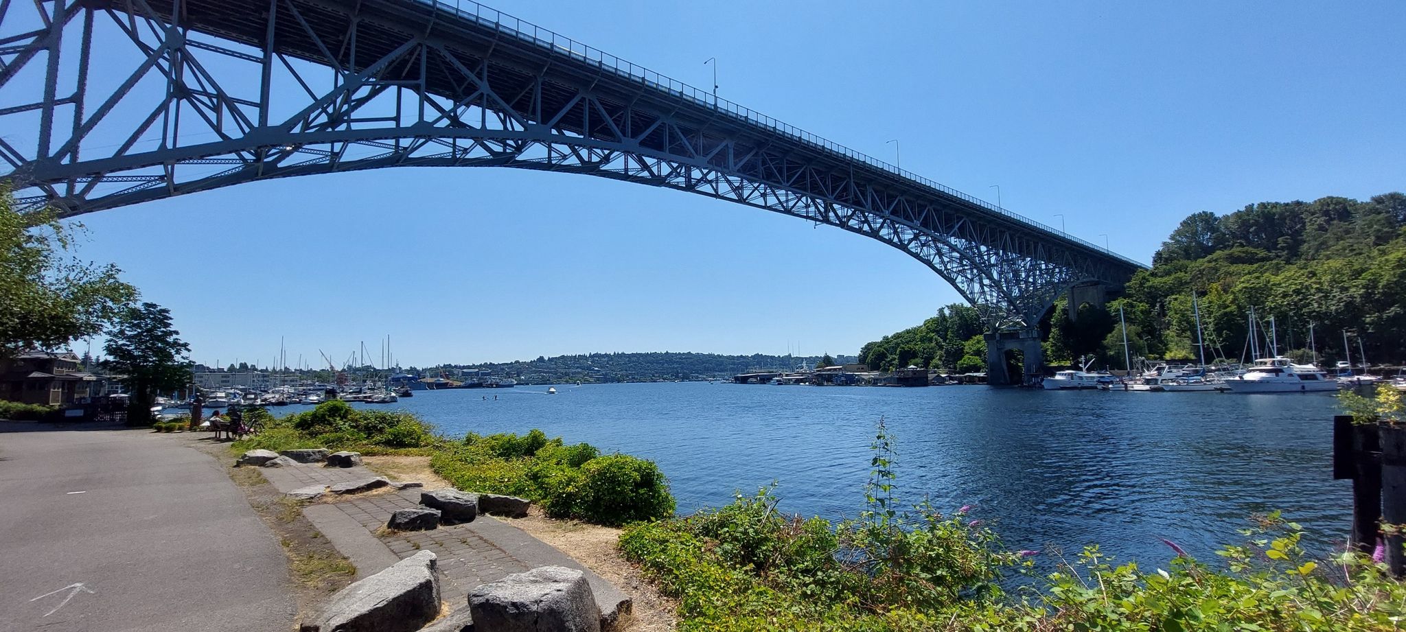 A large steel arch bridge spans a waterway with boats, under a clear blue sky. - Golden Garden – Salmon Life Cycle Sculpture loop from Canlis