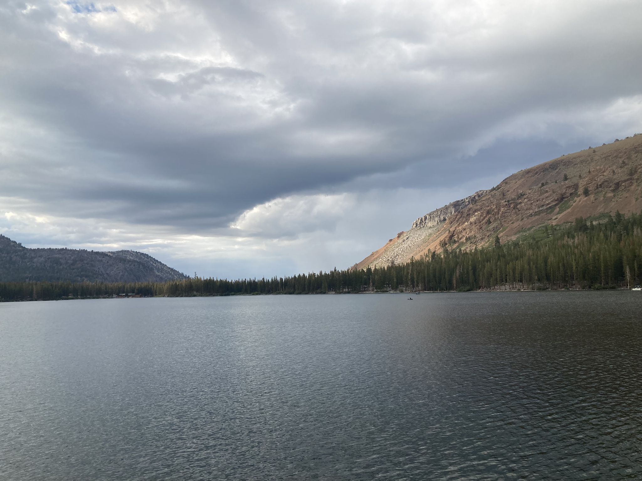 A wide, calm lake surrounded by forested hills under a dramatic cloudy sky. - Lakes Basin Path