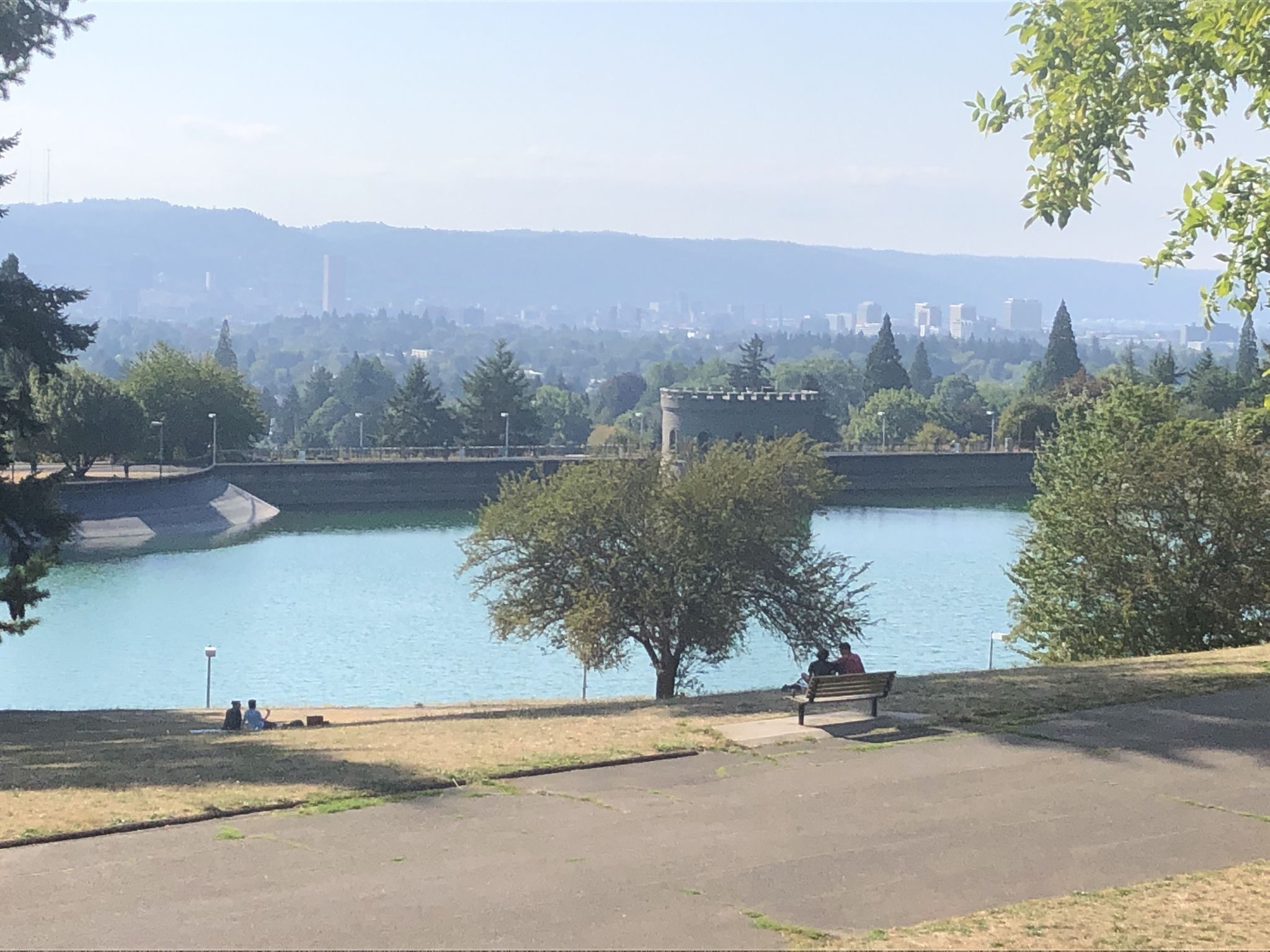 People relax by a reservoir with a distant city and hazy sky. - Blumenauer Bridge – Mount Tabor loop from Mount Hood Avenue