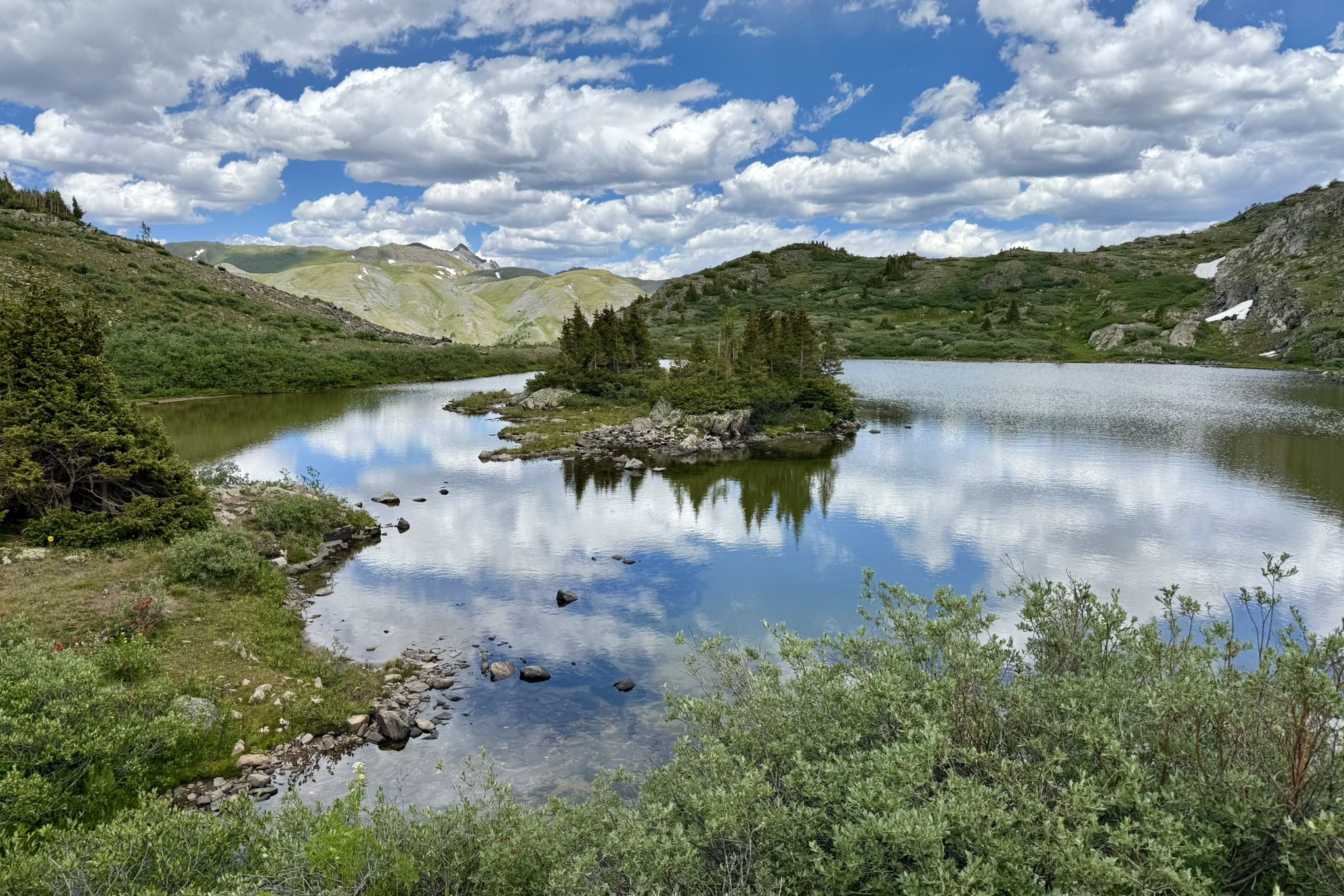 A lake with a small tree-covered island, reflecting clouds and mountains. - Highland Mary Lakes Loop Trail