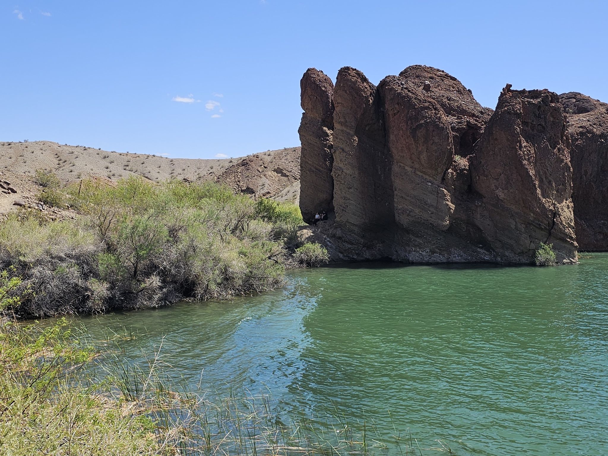 Green water next to large dark rock formations and desert shrubs. - SARA Trail loop