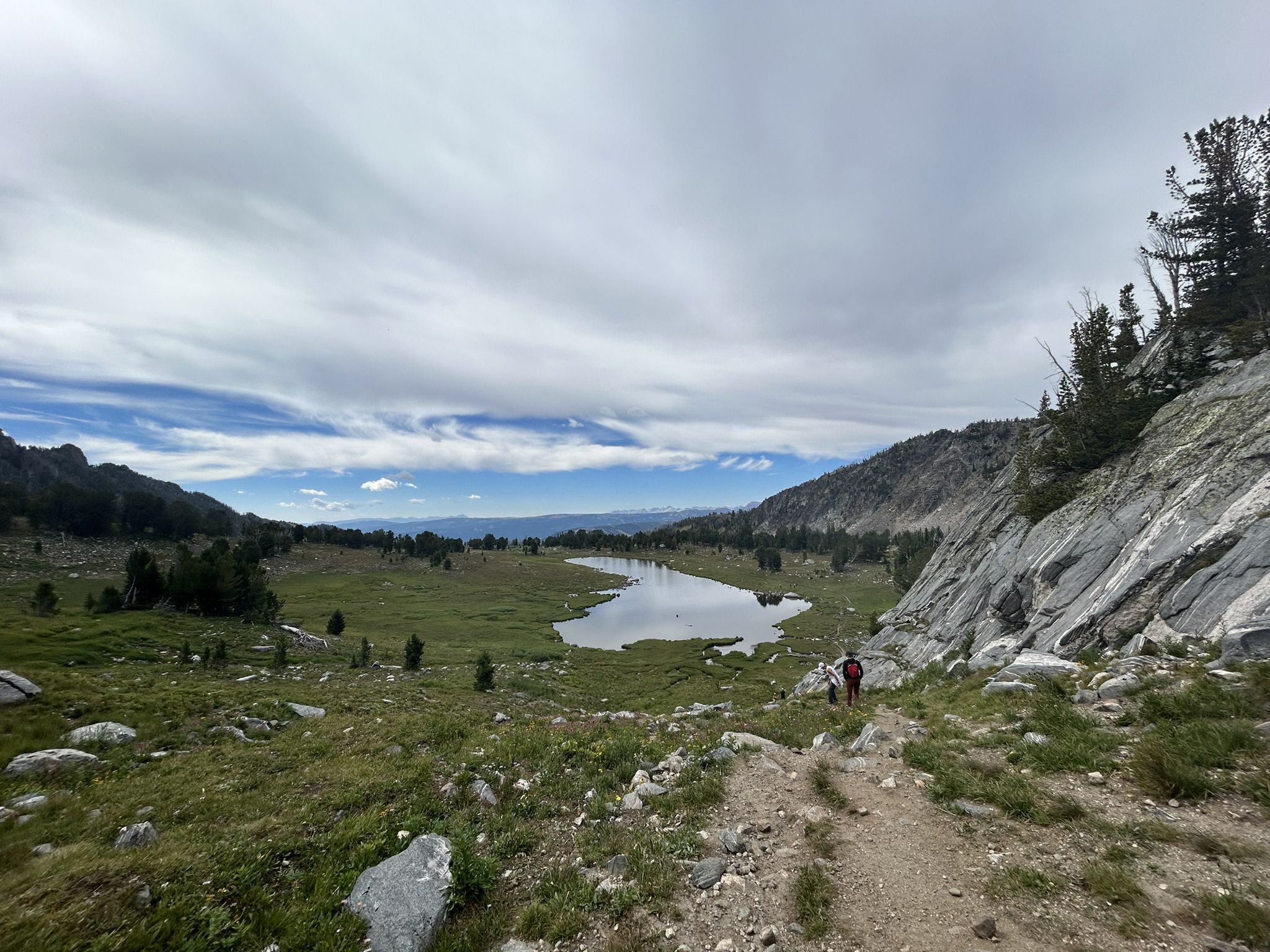 Hiker on a dirt path overlooking a lake in a green mountain valley. - Beehive Basin Trail No. 40