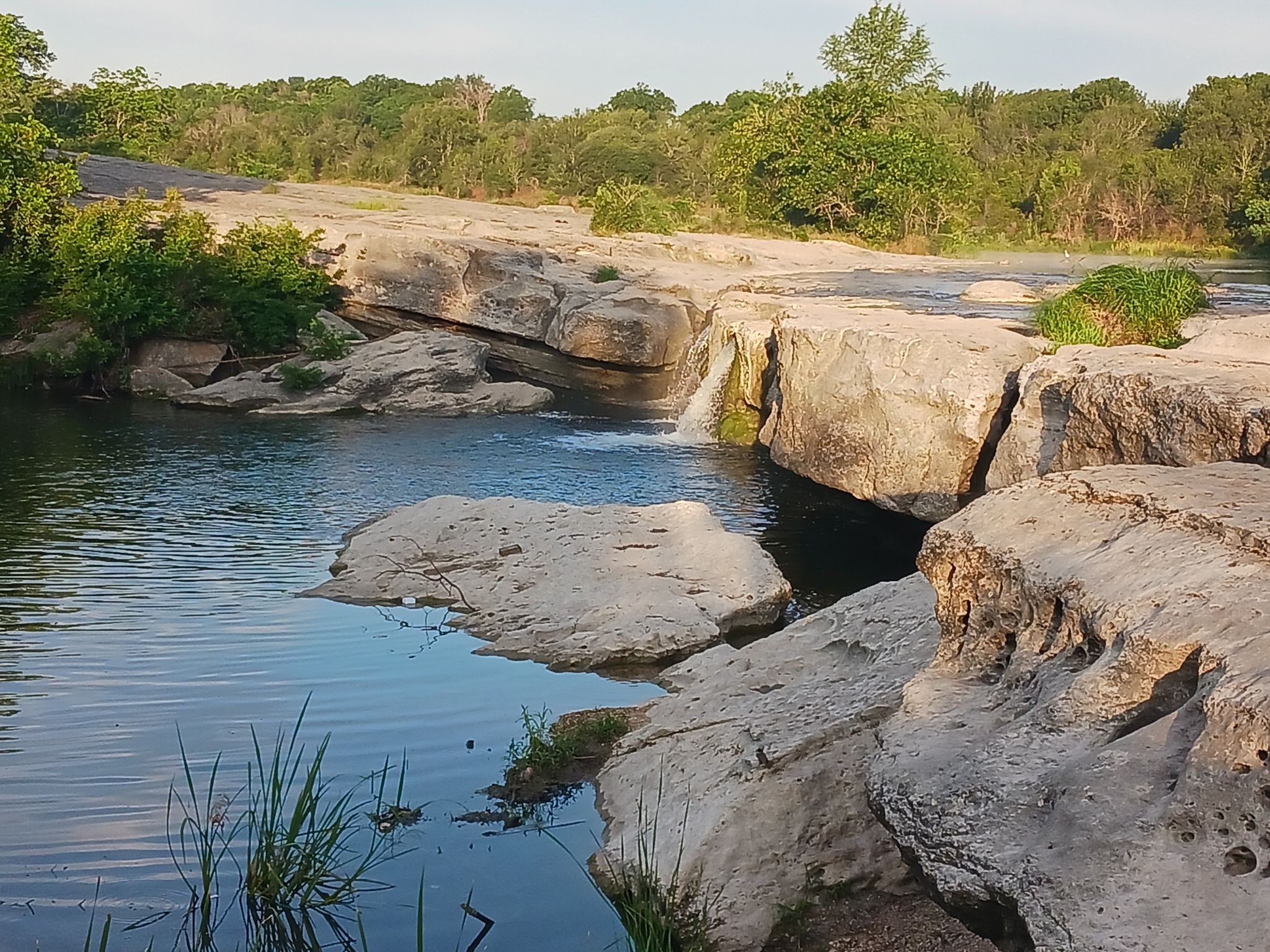 A river with rocky banks, a small waterfall, and green trees under bright sky. - Lower Falls and Homestead Loop Trail