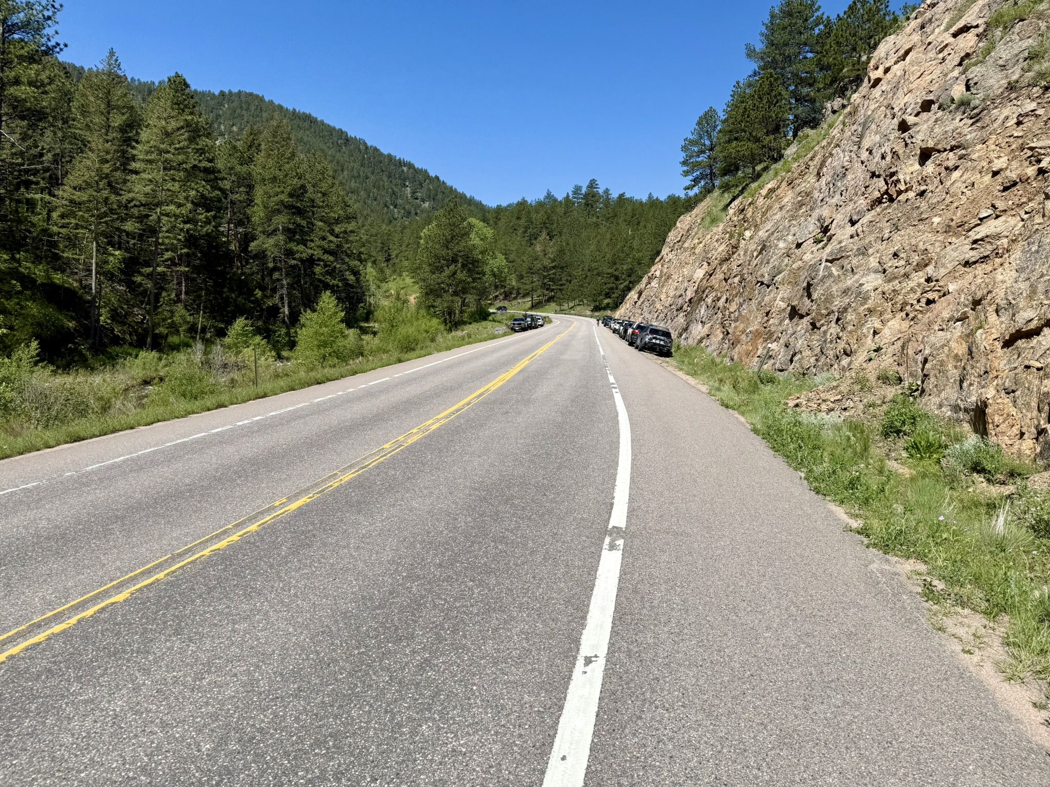 Straight road next to a rocky cliff and pine forest with parked cars. - Lefthand Trailhead loop from Buckingham Park