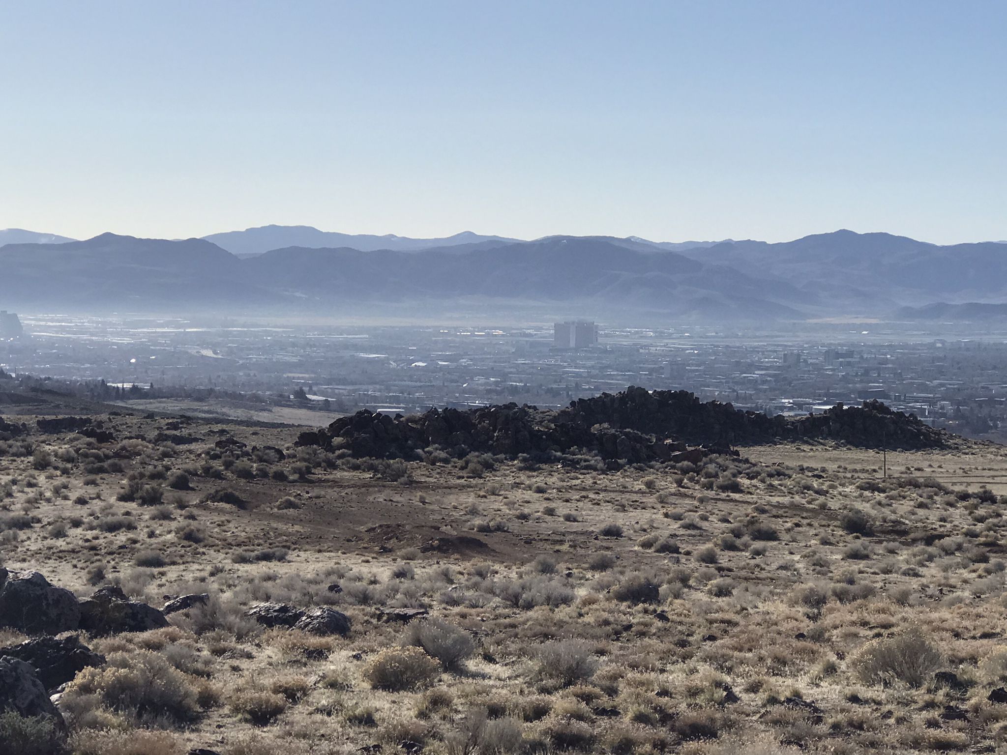 Panoramic view of a hazy city in a valley, surrounded by distant mountains. - Bench with a view – Poedunk Trail loop from University of Nevada, Reno