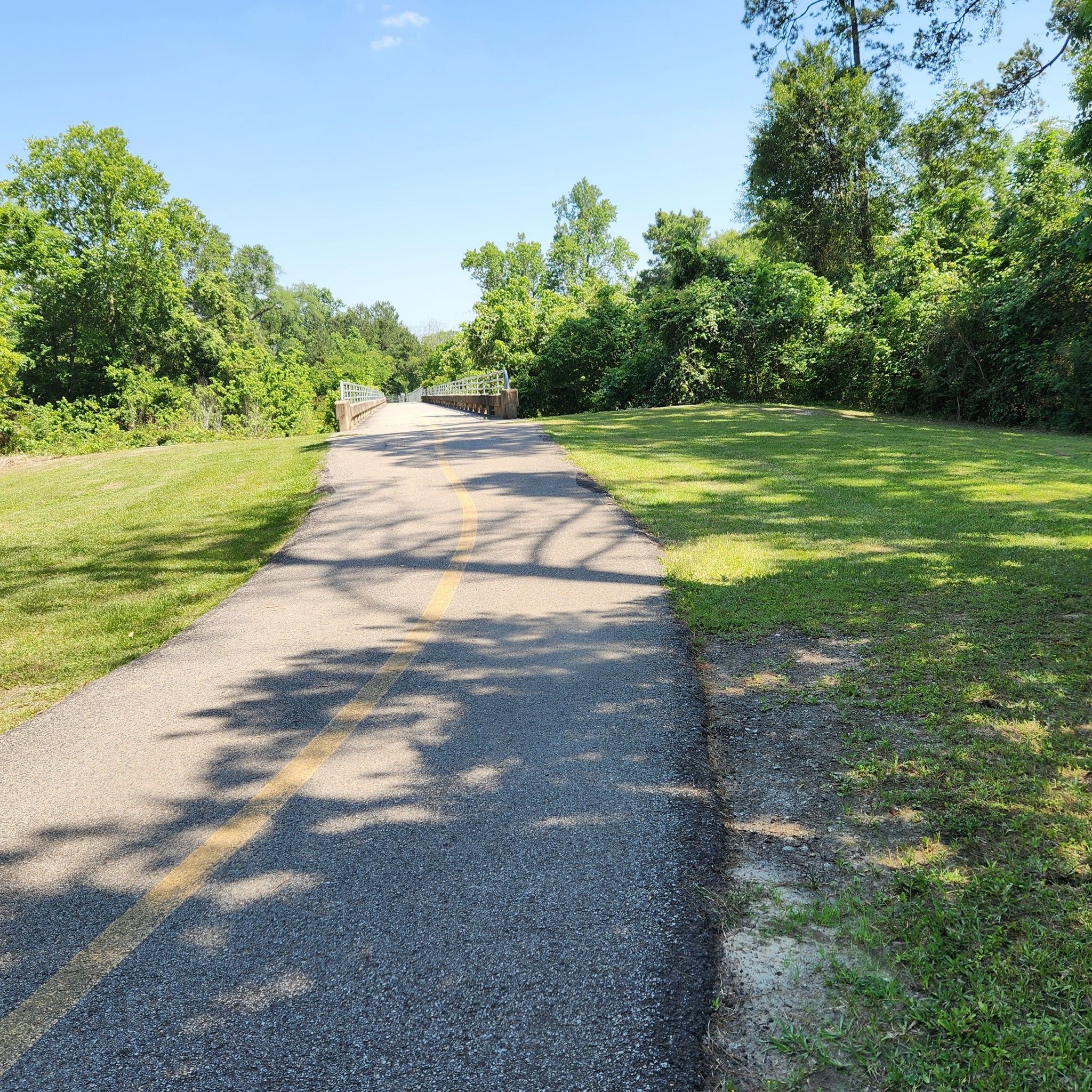 Paved path with yellow line leading to a bridge, surrounded by green grass. - Spring Creek Greenway to Jesse Jones Park