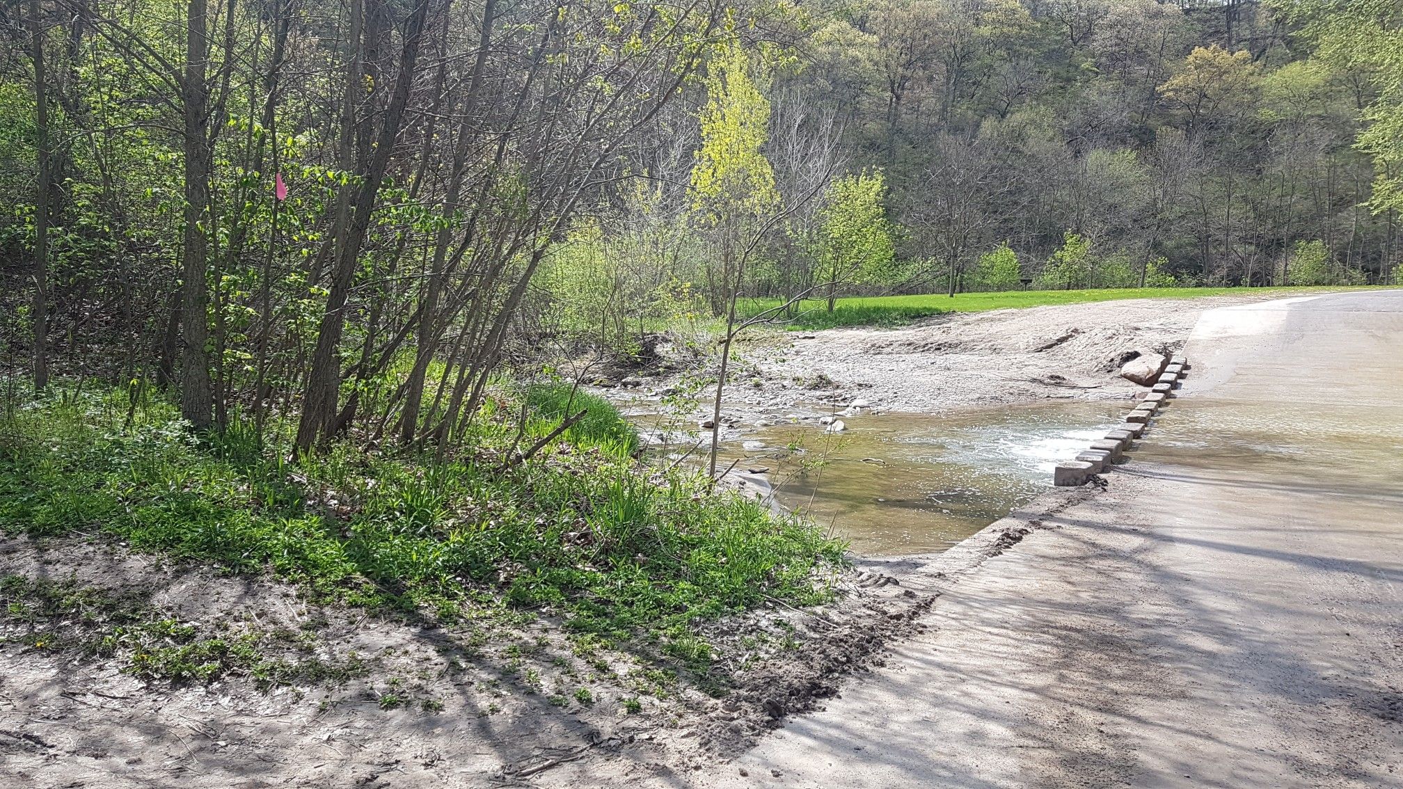 Sunny view of a stream flowing across a dirt road in a wooded area. - Stone bridge – Peas Creek crossing loop from Ledges State Park
