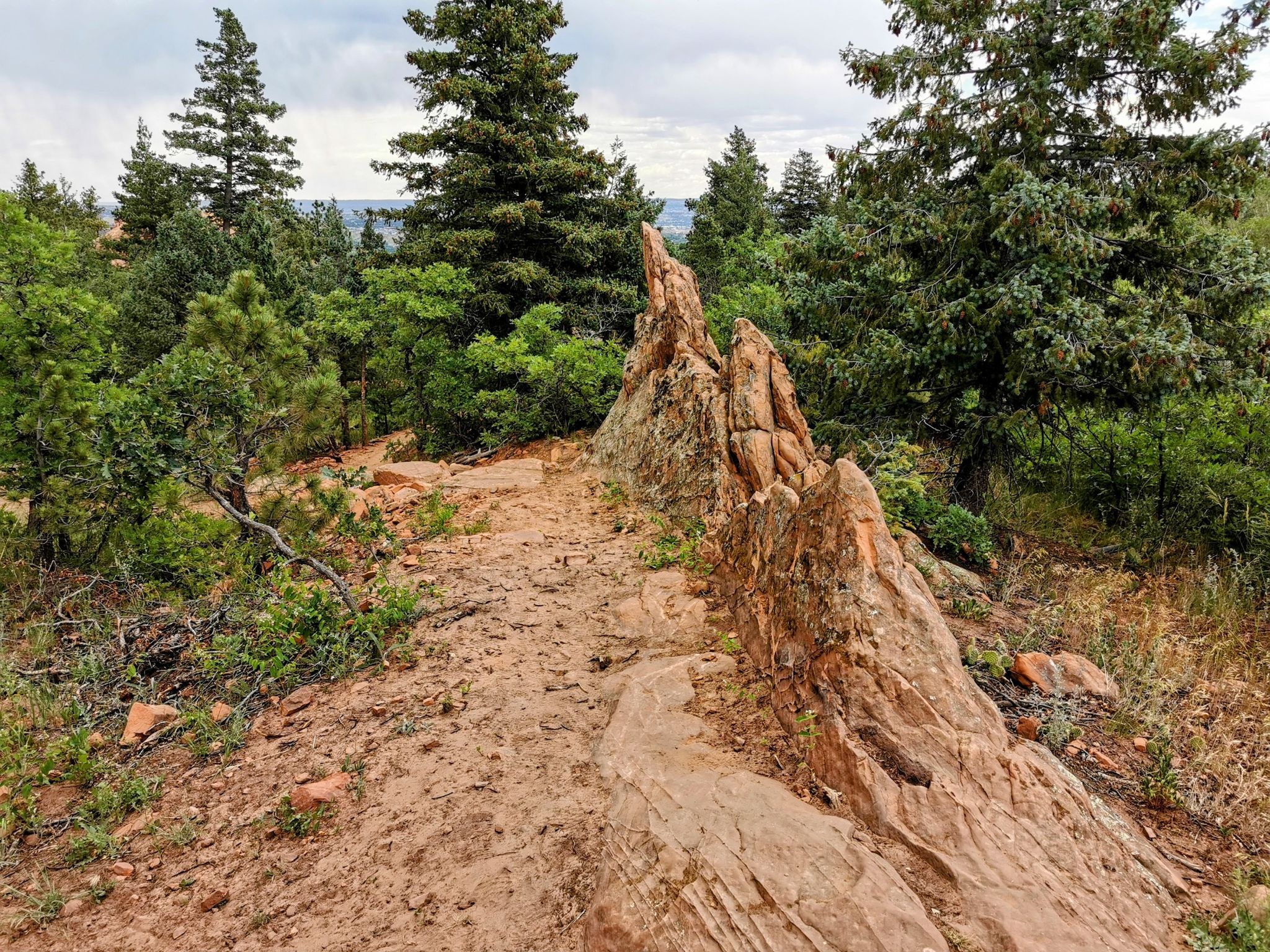 A jagged rock formation beside a dirt path through a green forest. - Intemann Trail