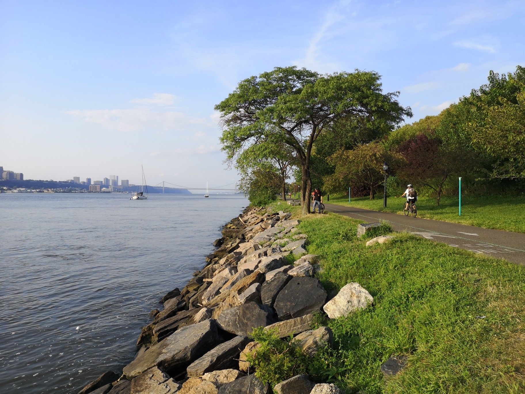 Cyclists on a path beside a rocky riverbank with a distant city. - Hudson River Greenway – Hudson River Greenway loop from 145th Street
