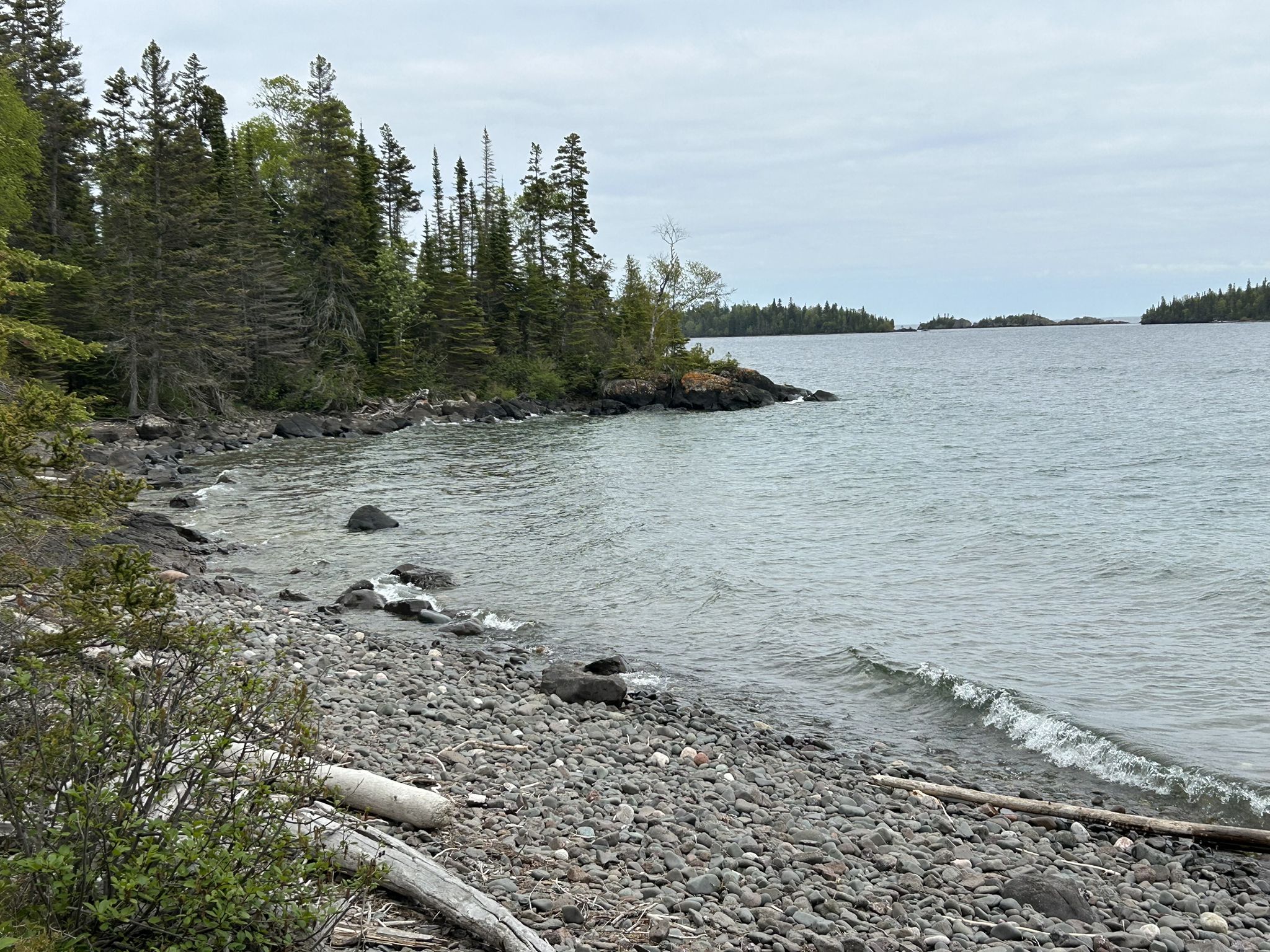 Rocky beach with small waves, evergreen trees, and distant islands under a cloudy sky. - Rock Harbor Trail