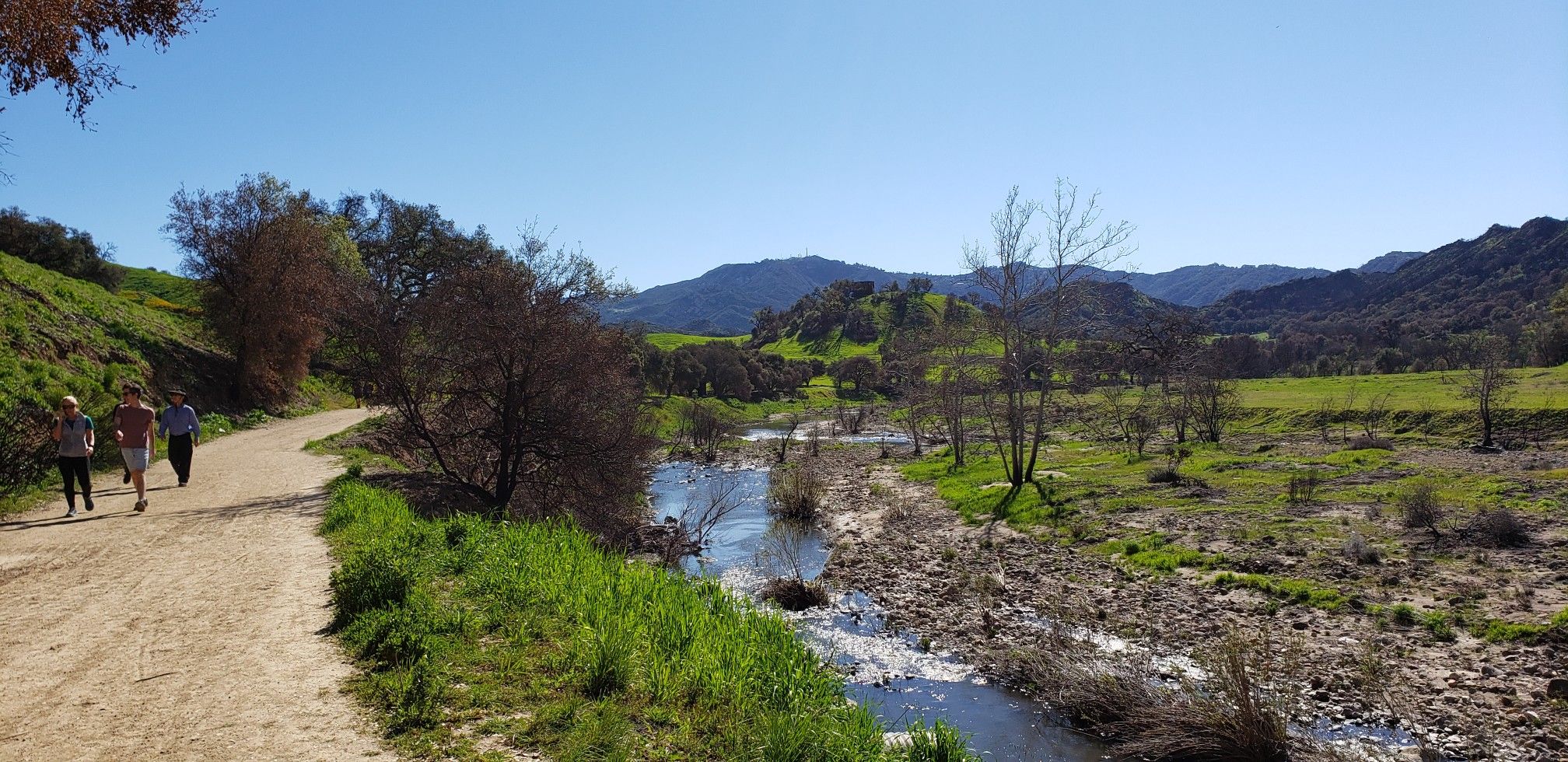 People walking on a dirt path beside a river in a green valley. - Grasslands Trailhead – Las Virgenes Fire Road Trail loop from Las Virgenes MWD Open Space