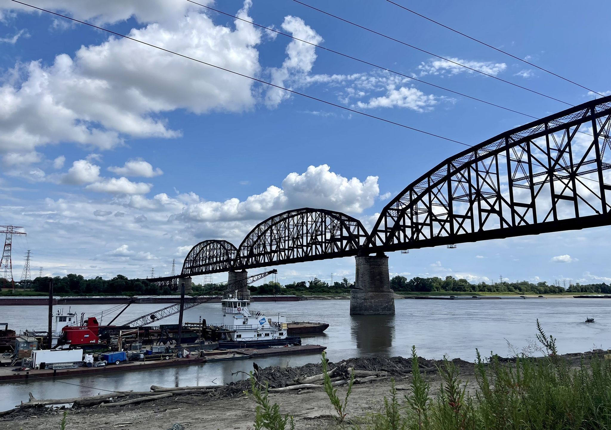 An arched metal truss bridge spans a river with barges docked below. - Riverfront Trail – Old Courthouse loop from Central West End