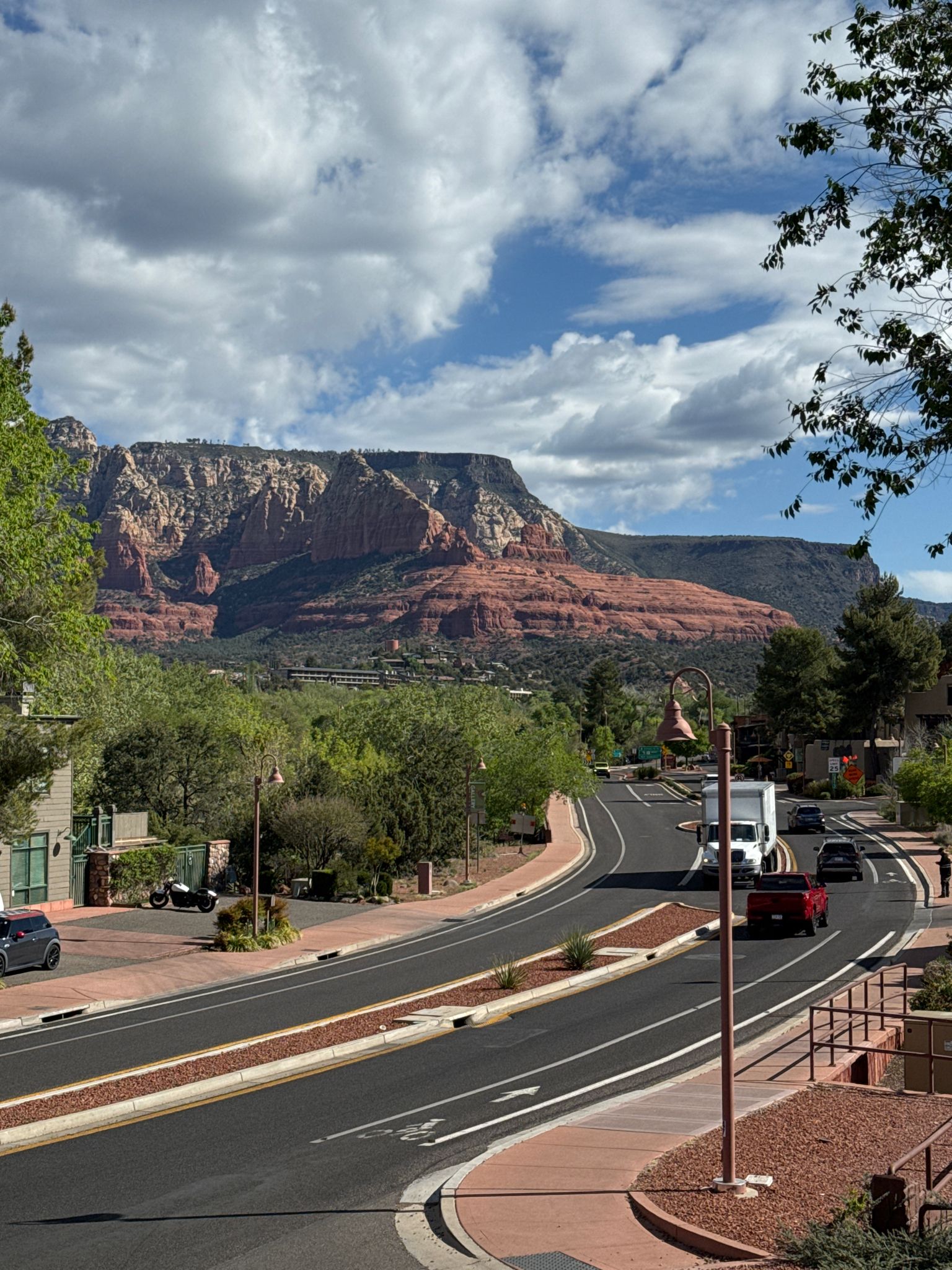 Winding road with cars, green trees, and large red rock mountains under cloudy sky. - Chapel of the Holy Cross via Red Rock Scenic Byway