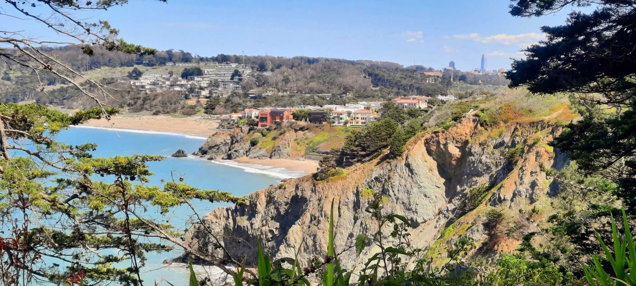 Panoramic view of a rocky coastline, sandy beach, and distant buildings under blue sky. - Coastal Trail