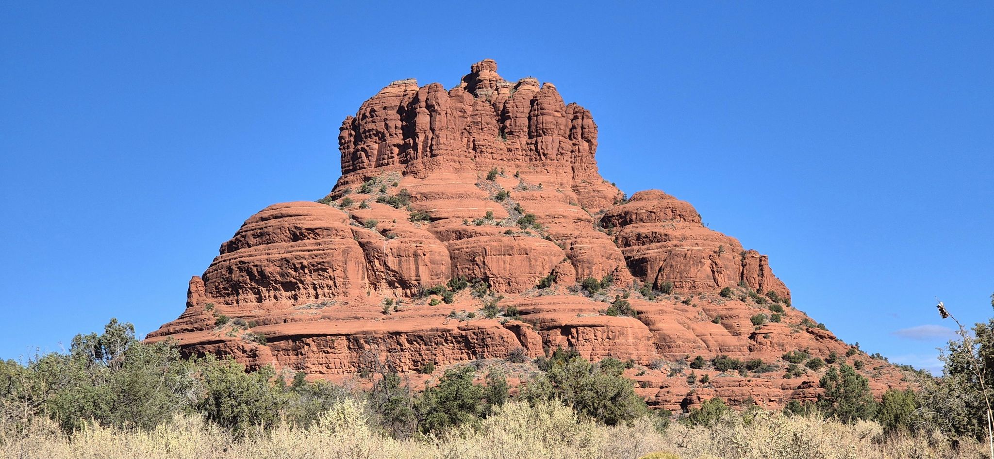 Massive red rock formation with distinct layers, surrounded by low brush. - Bell Rock Trail