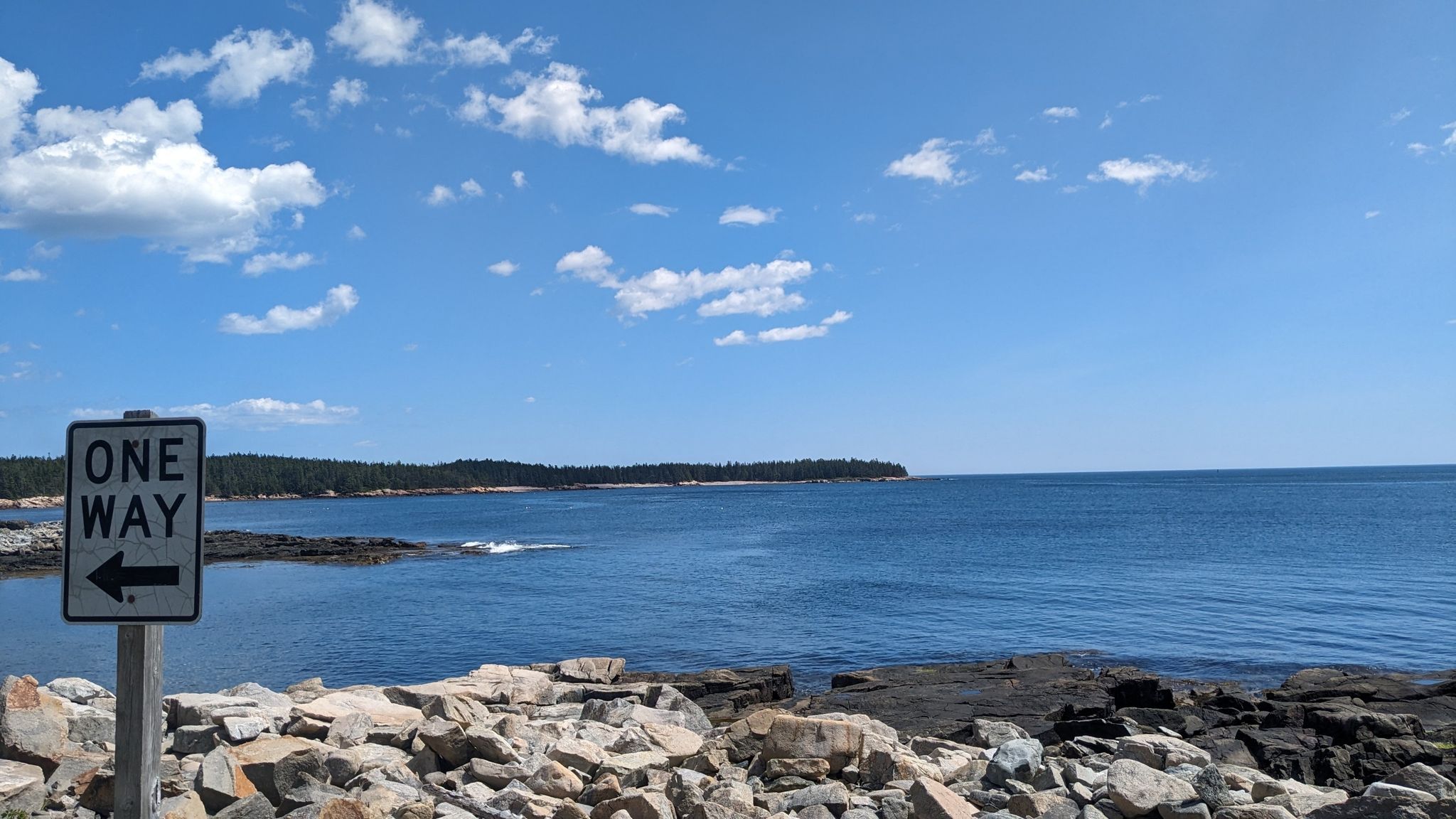 A "One Way" sign overlooks a rocky coastline and blue ocean on a sunny day. - Birch Harbor and Buck Cove Loop