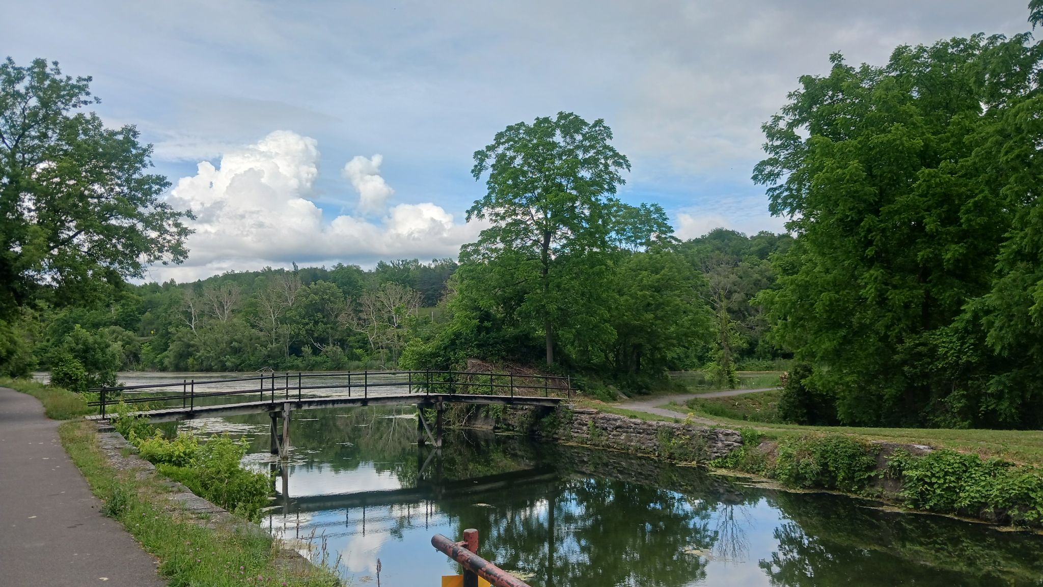Canal with a small bridge, surrounded by green trees under a cloudy sky. - Canal Landing to Erie Canalway Trail