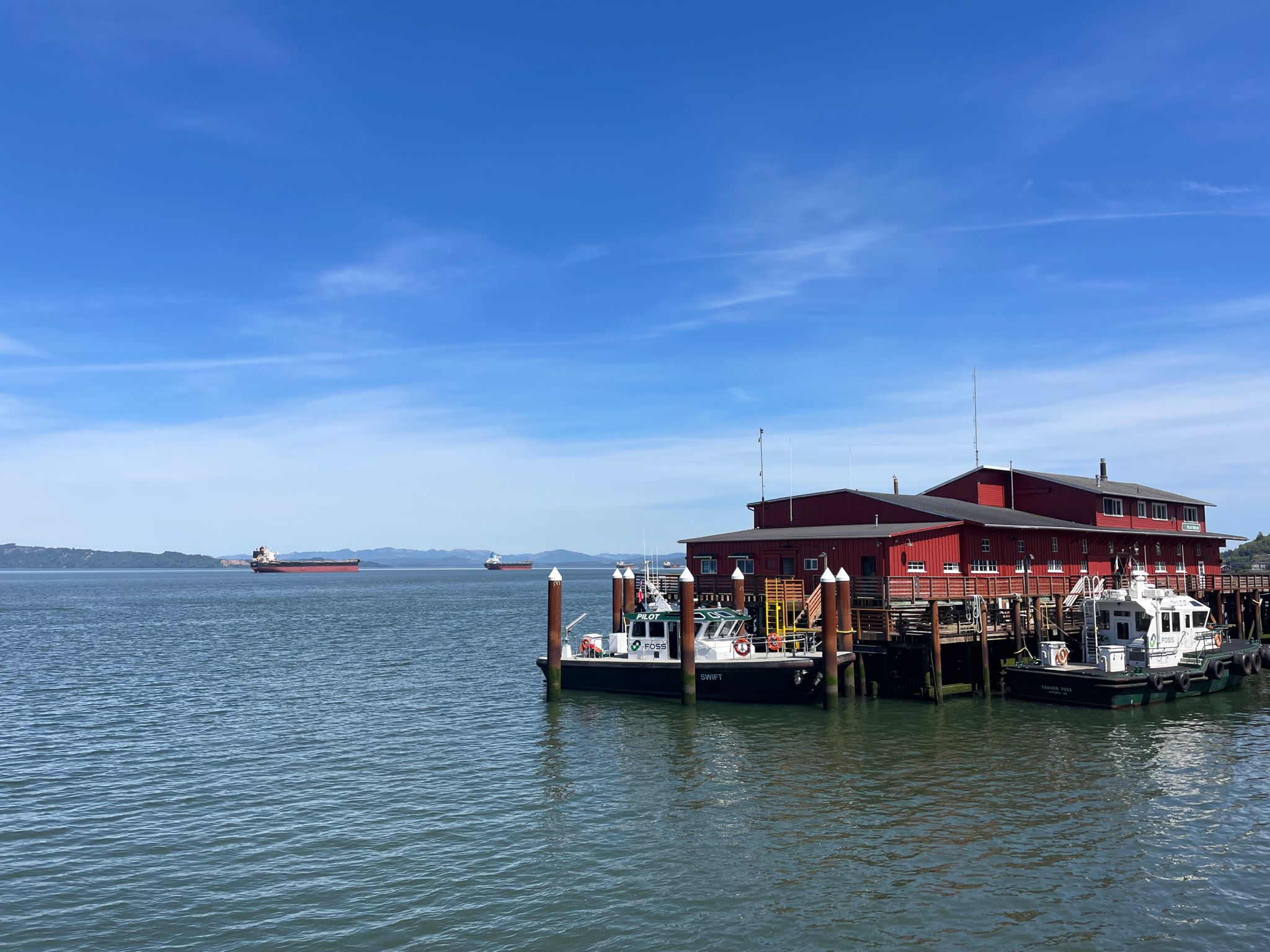Red building on a pier with boats, calm water, distant ships, clear blue sky. - Youngs River loop