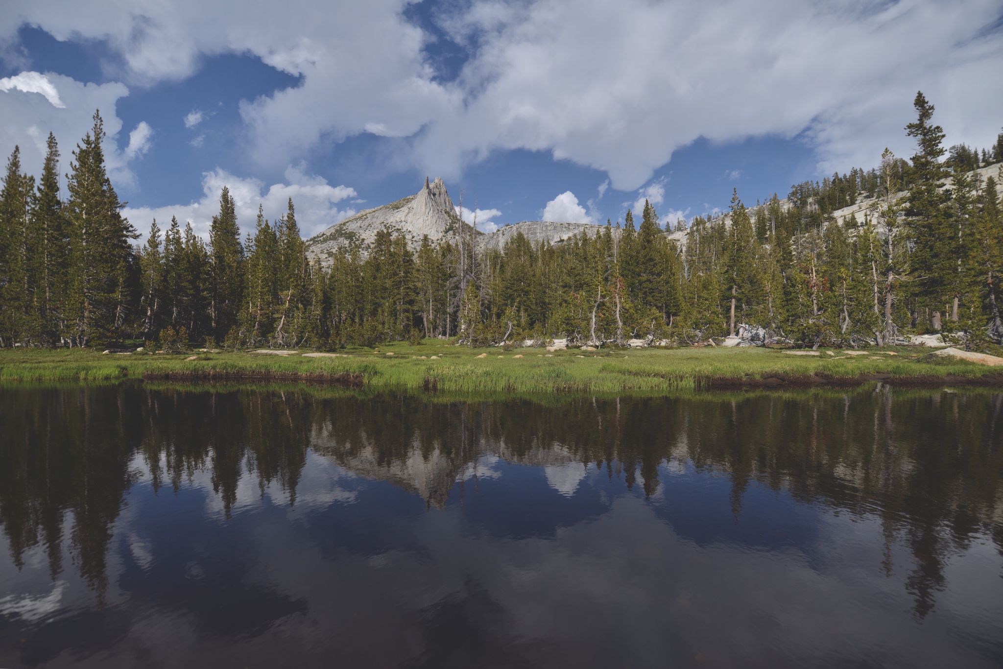 A calm lake reflects pine trees and a distant rocky mountain peak. - Upper Cathedral Lake – John Muir Trail loop from Tuolumne Meadows