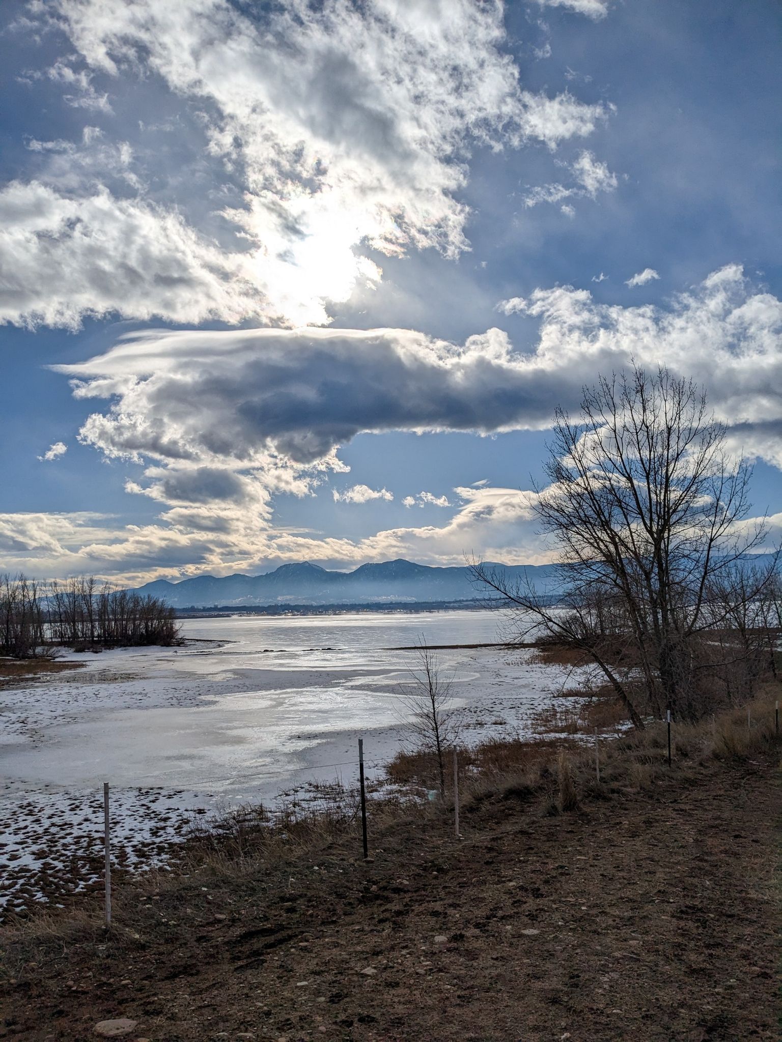 Partially frozen lake with bare trees and mountains under a bright cloudy sky. - Boulder Reservoir Loop Trail