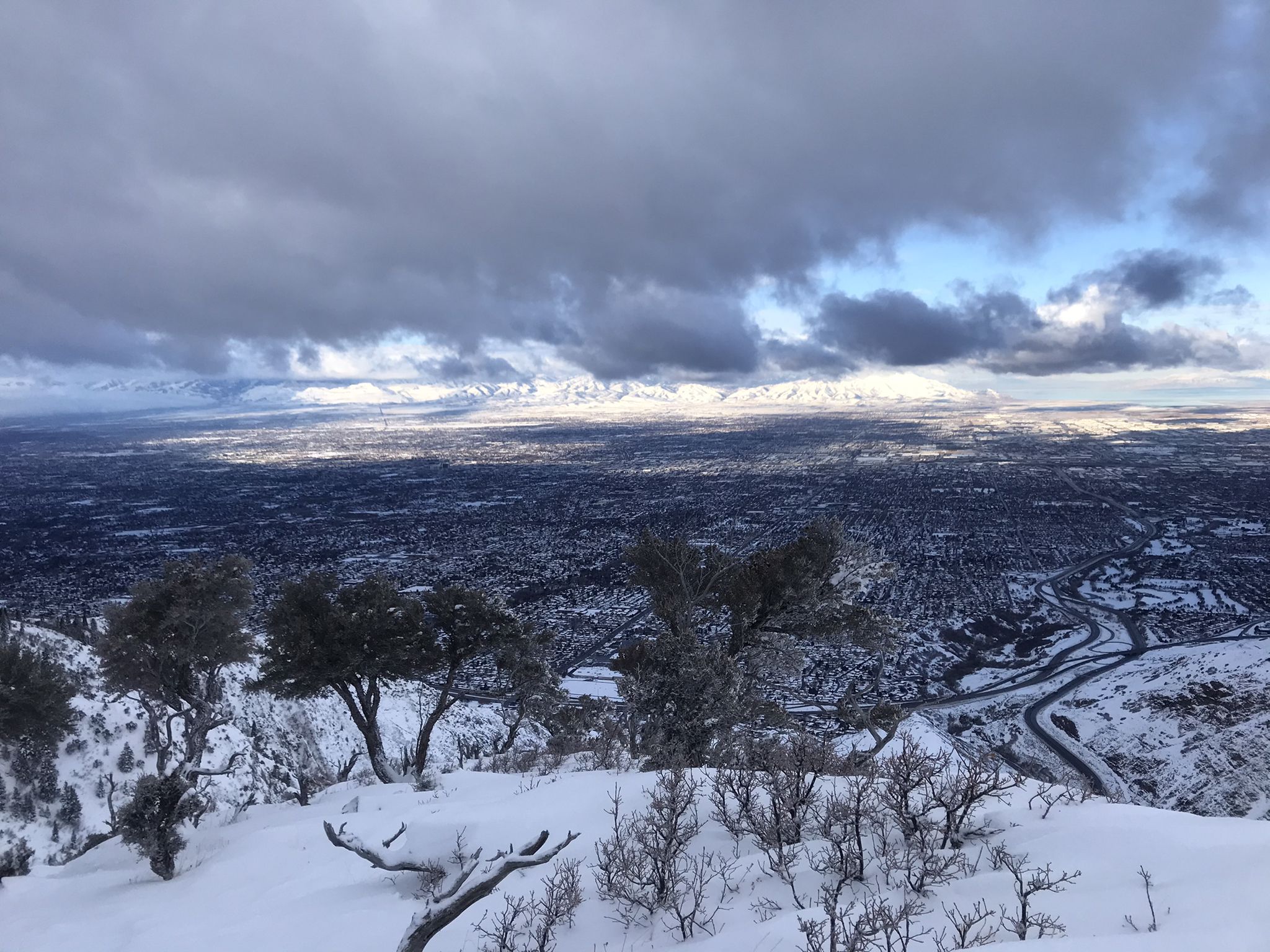 Snowy mountain view overlooking a city under dramatic, cloudy skies. - Grandeur Peak Loop